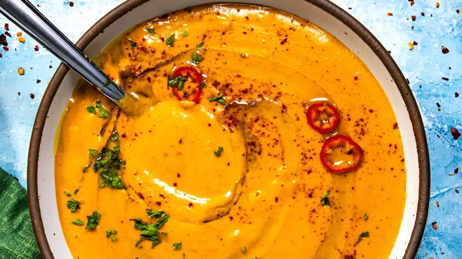 A bowl of creamy orange soup garnished with sliced red chili, chopped herbs, and a sprinkle of spices, with a spoon resting in the bowl.
