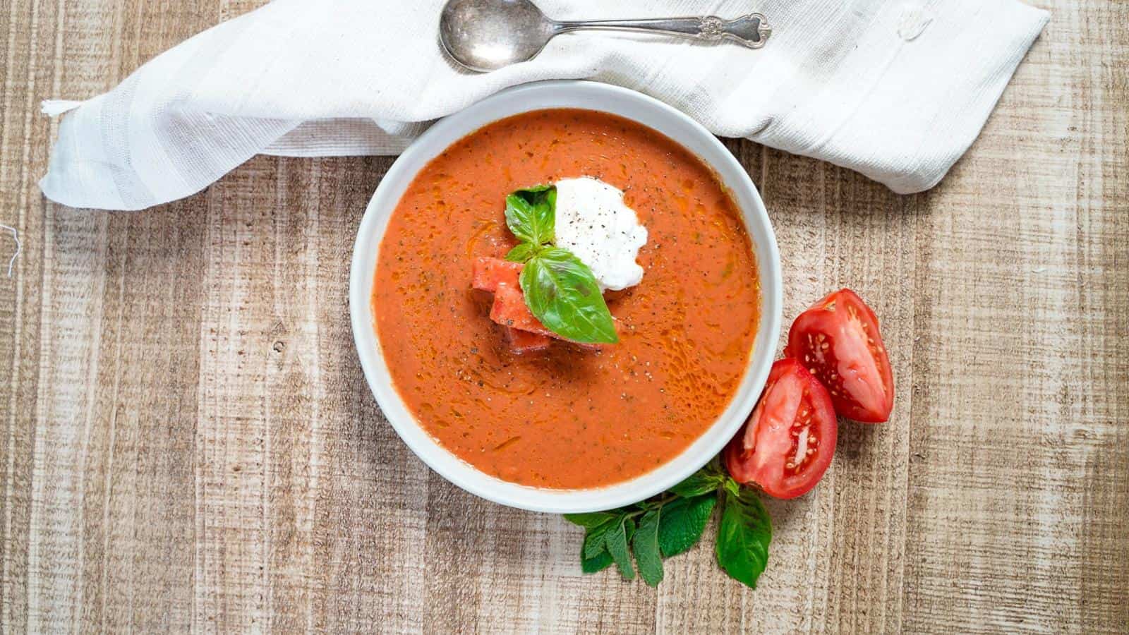 A bowl of tomato soup garnished with cream and basil, placed on a wooden table with a cloth napkin, spoon, and sliced tomatoes on the side.