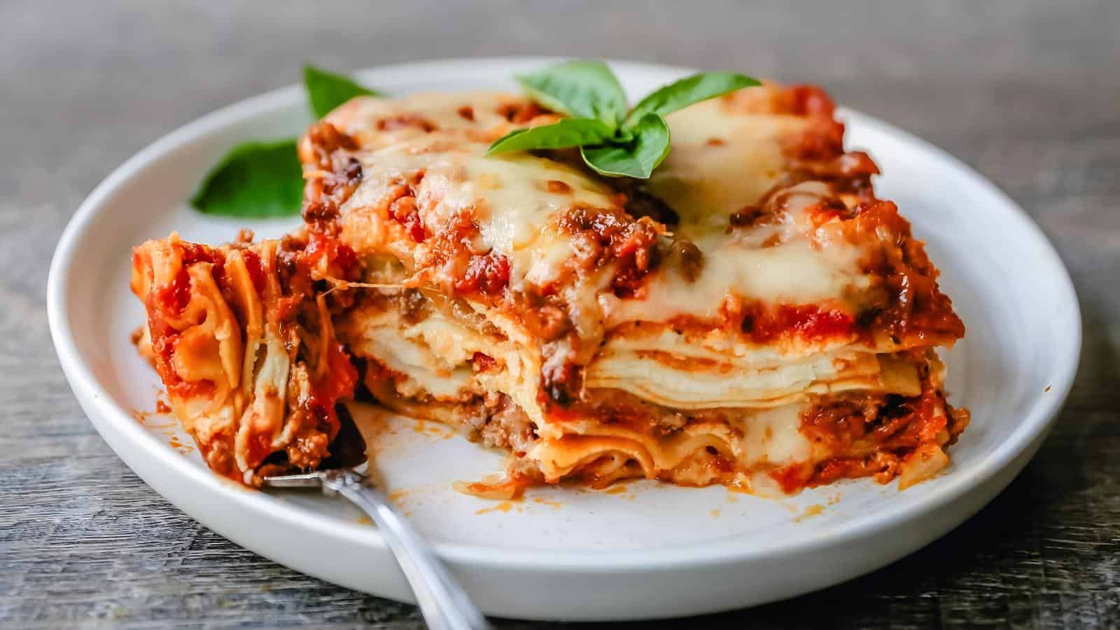 A slice of lasagna with layers of pasta, meat sauce, and melted cheese, garnished with fresh basil, on a white plate with a fork.