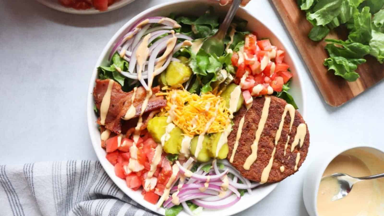 A salad bowl with lettuce, tomato, red onion, pickles, shredded cheese, bacon, a burger patty, and dressing, viewed from above.