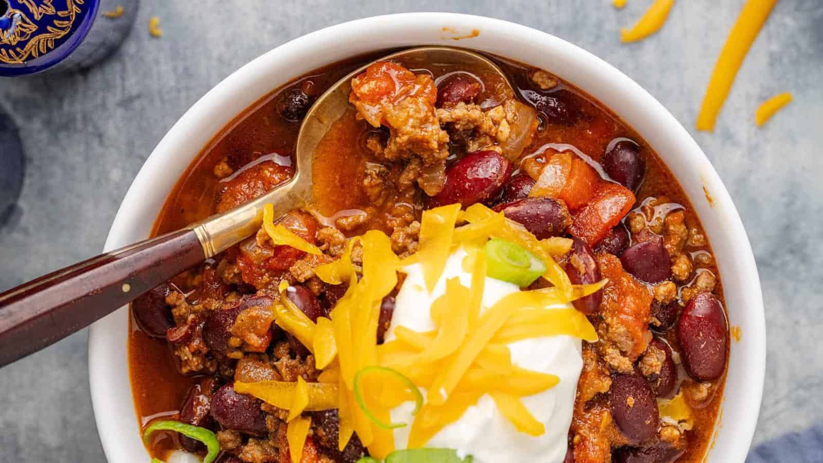 A bowl of chili with ground beef, beans, diced tomatoes, shredded cheddar cheese, sour cream, and sliced green onions, with a spoon resting in the bowl.