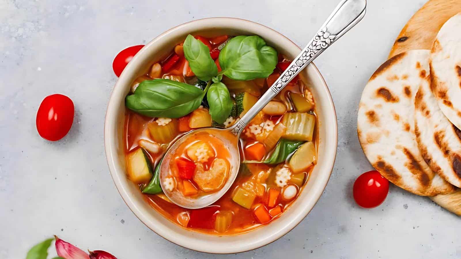 A bowl of vegetable soup with a spoon, topped with fresh basil, surrounded by cherry tomatoes and flatbread slices on a light surface.
