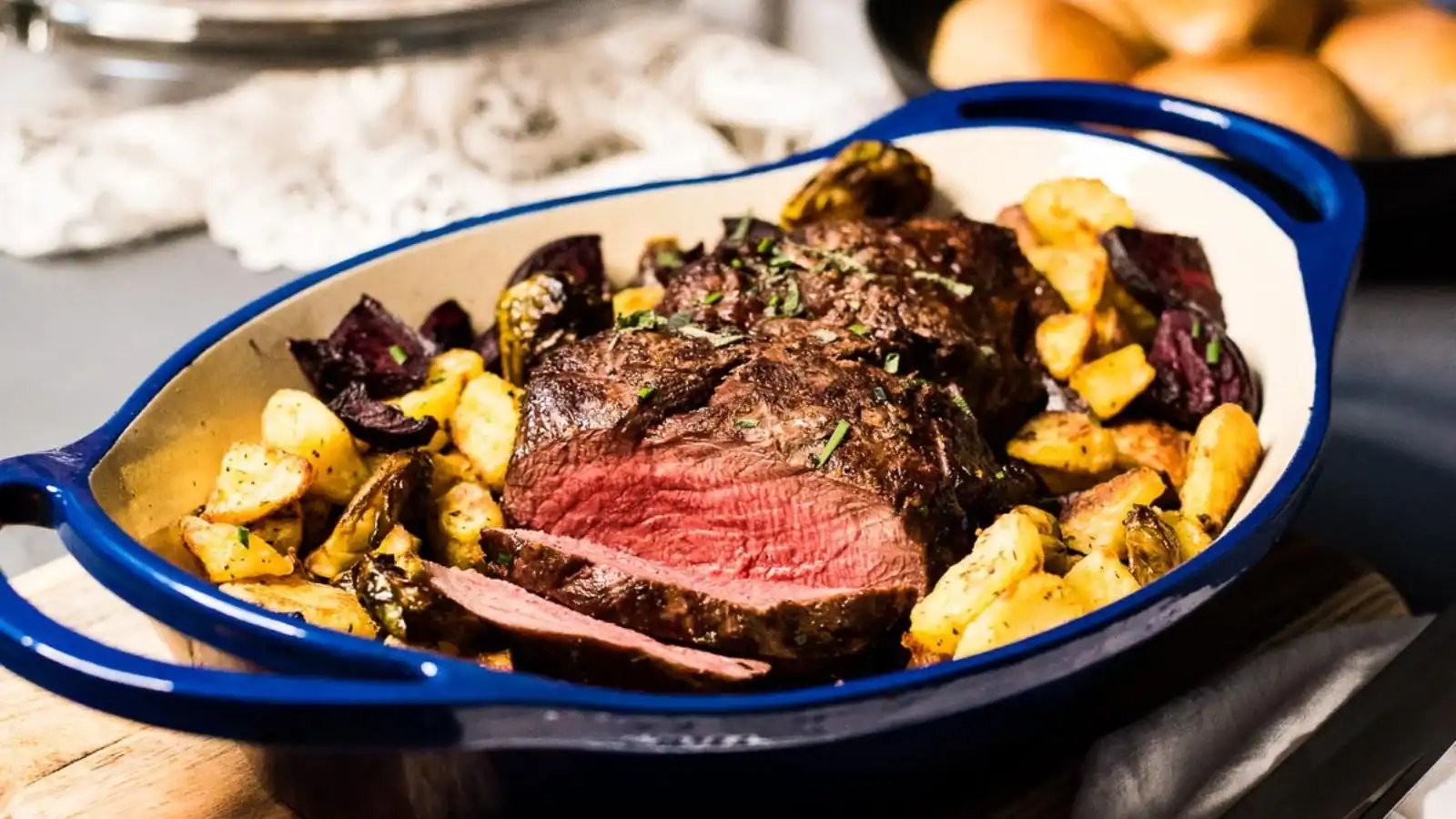 A blue roasting dish with sliced roast beef, surrounded by roasted potatoes and vegetables, sits on a wooden board.