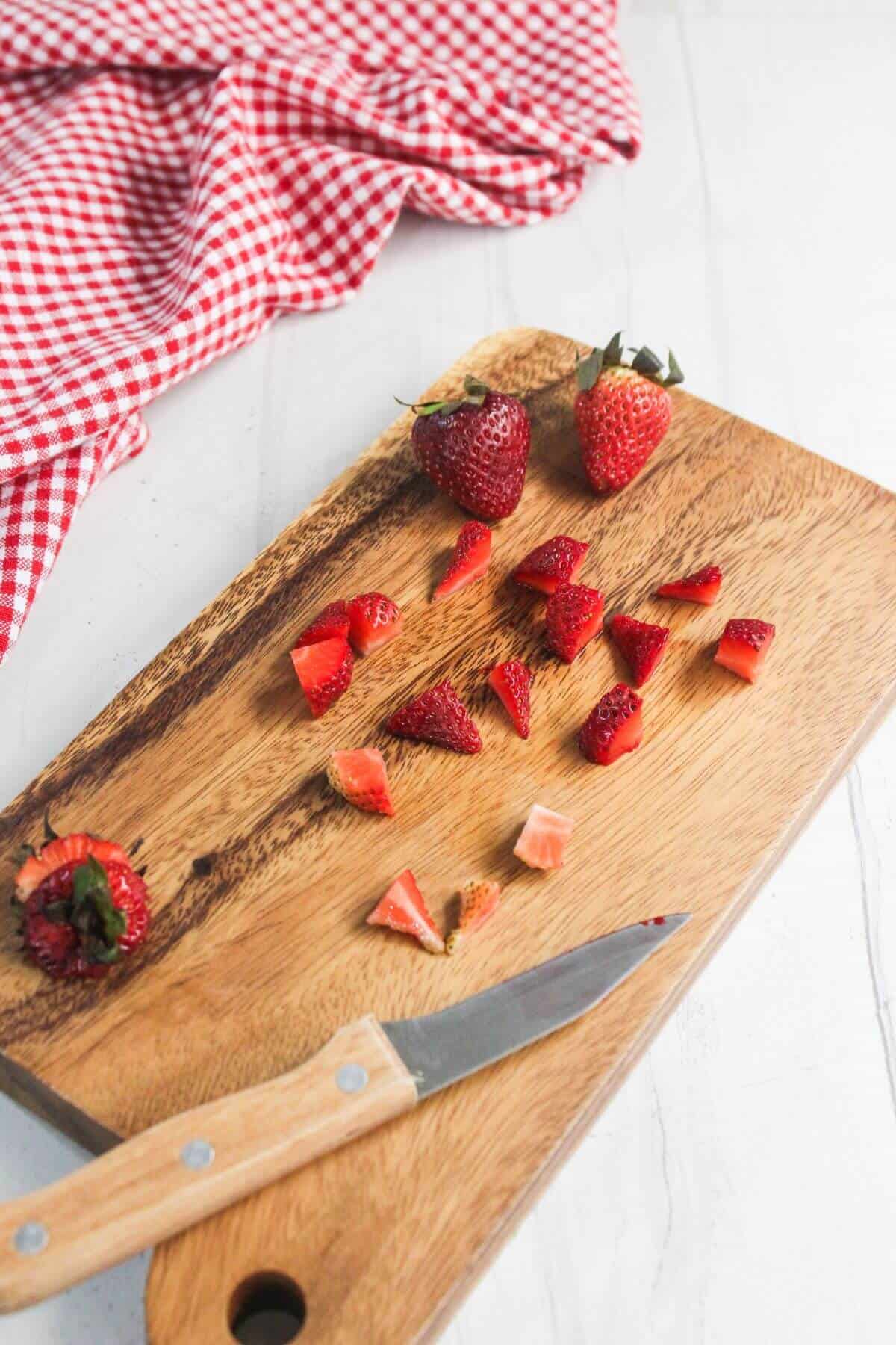 A wooden cutting board with a paring knife, chopped strawberries, and a red gingham cloth in the background.