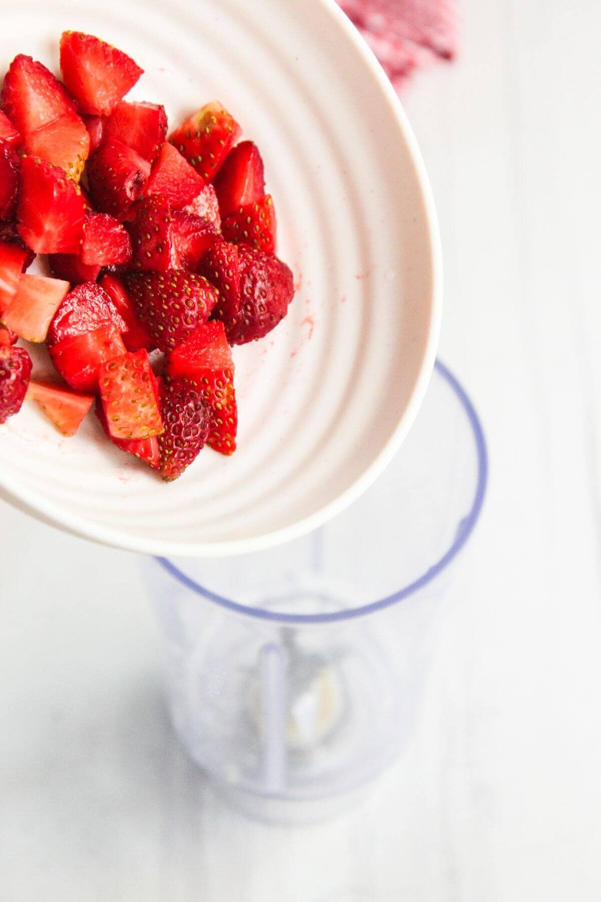 Chopped strawberries are being poured from a white plate into a blender on a white surface.