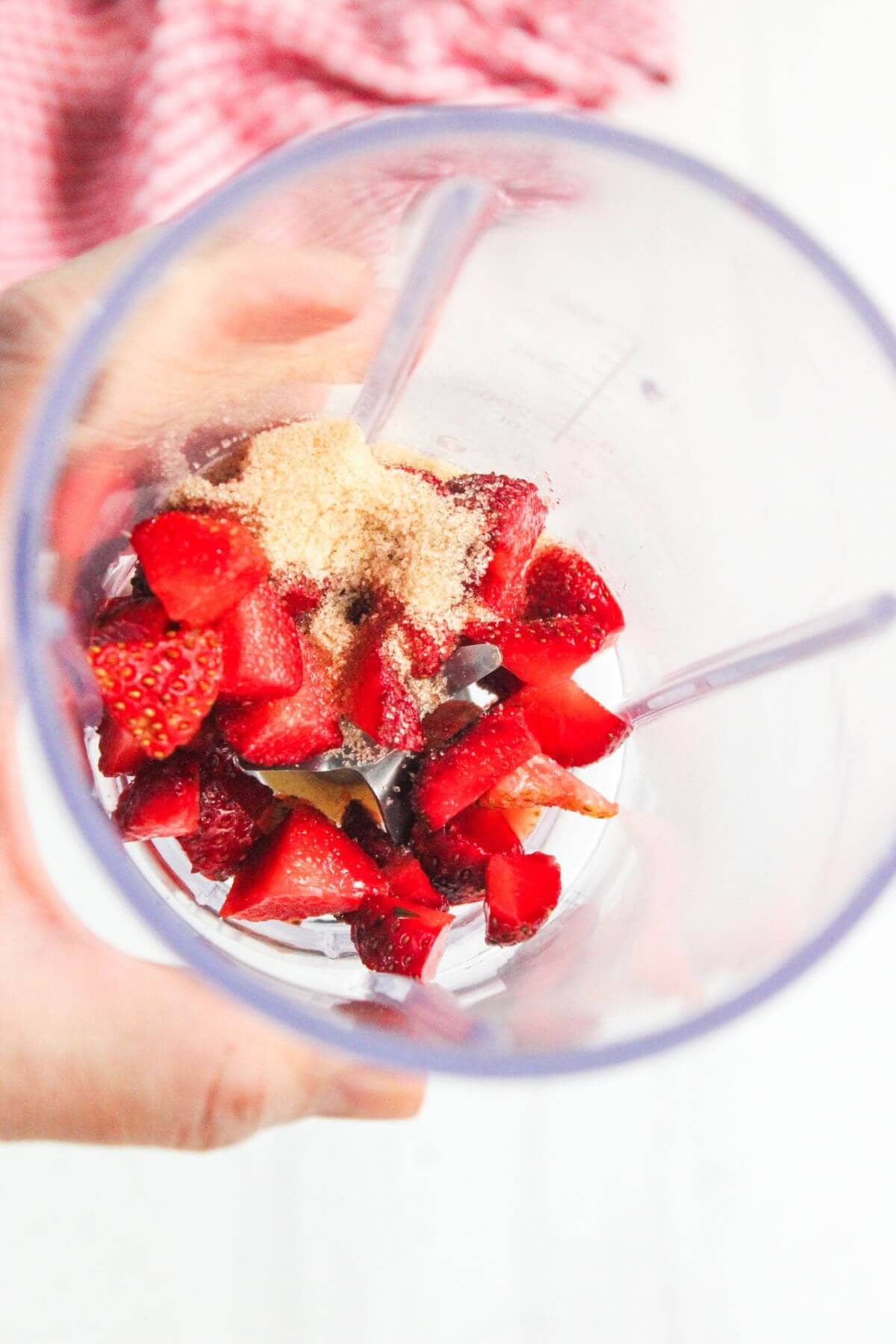 Overhead view of a blender cup containing chopped strawberries, brown sugar, and other ingredients, held by a hand.