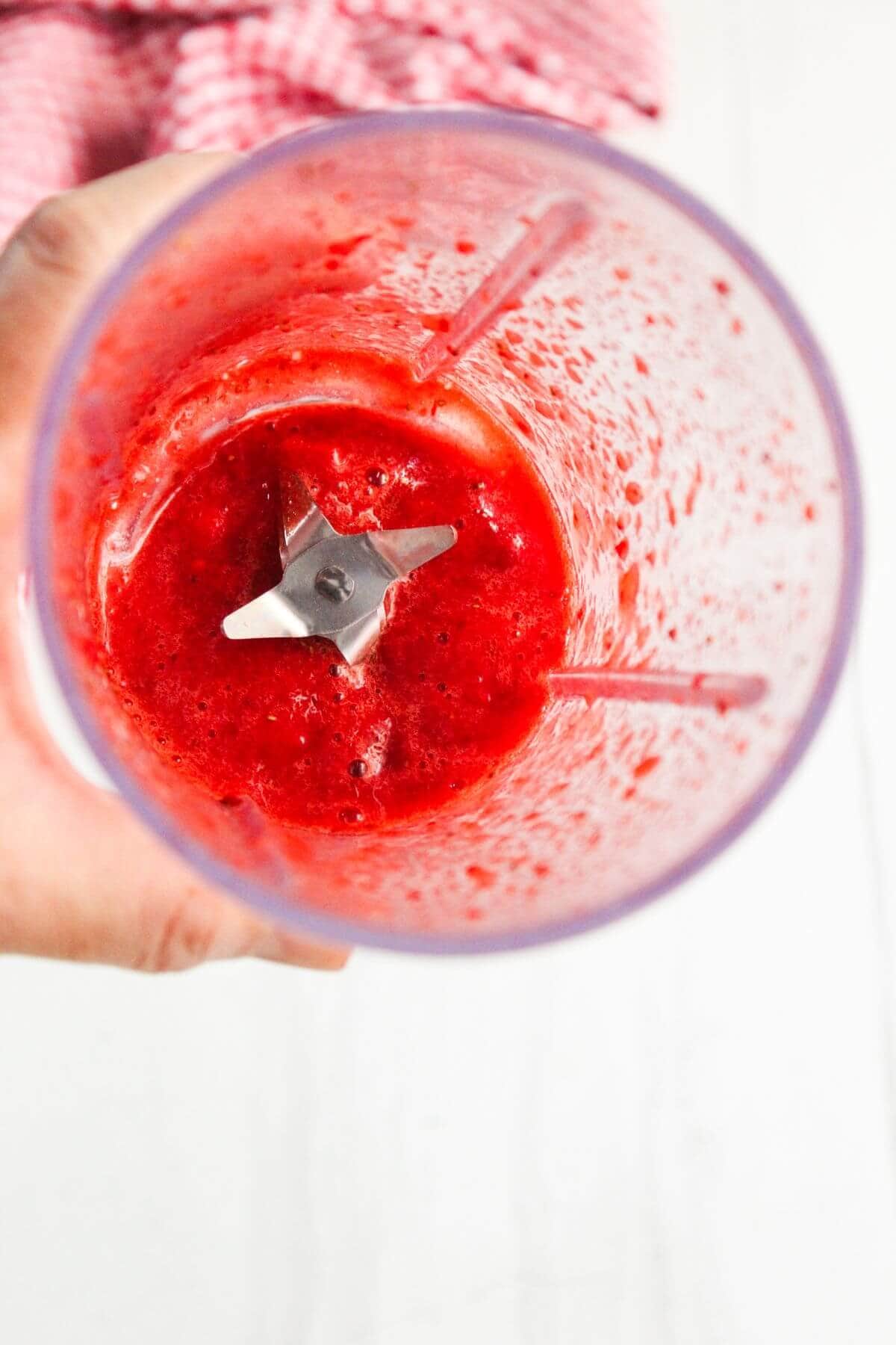 A top-down view of a blender containing a red fruit puree, with visible blades and a textured pink-red mixture inside.
