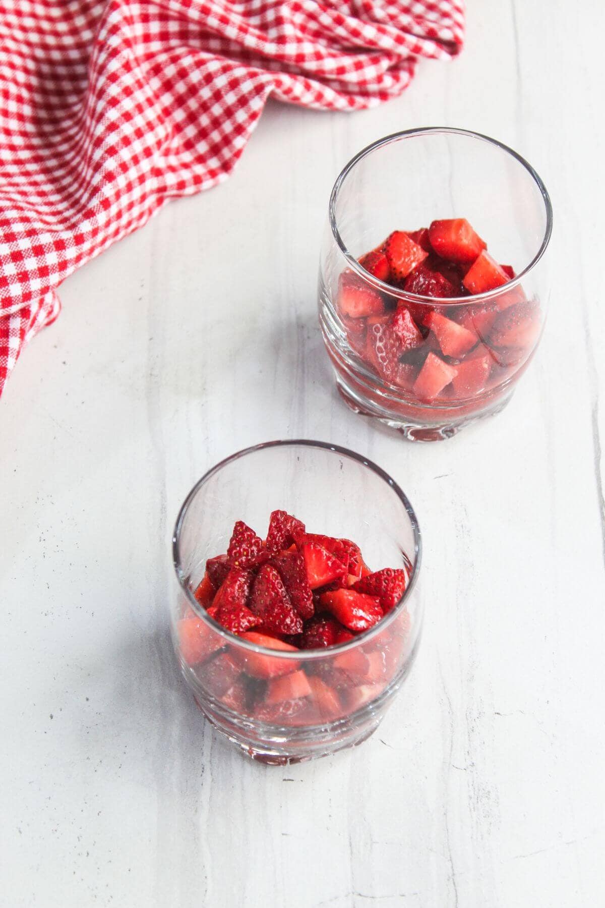 Two clear glasses filled with chopped strawberries on a white surface, with a red and white checkered cloth in the background.