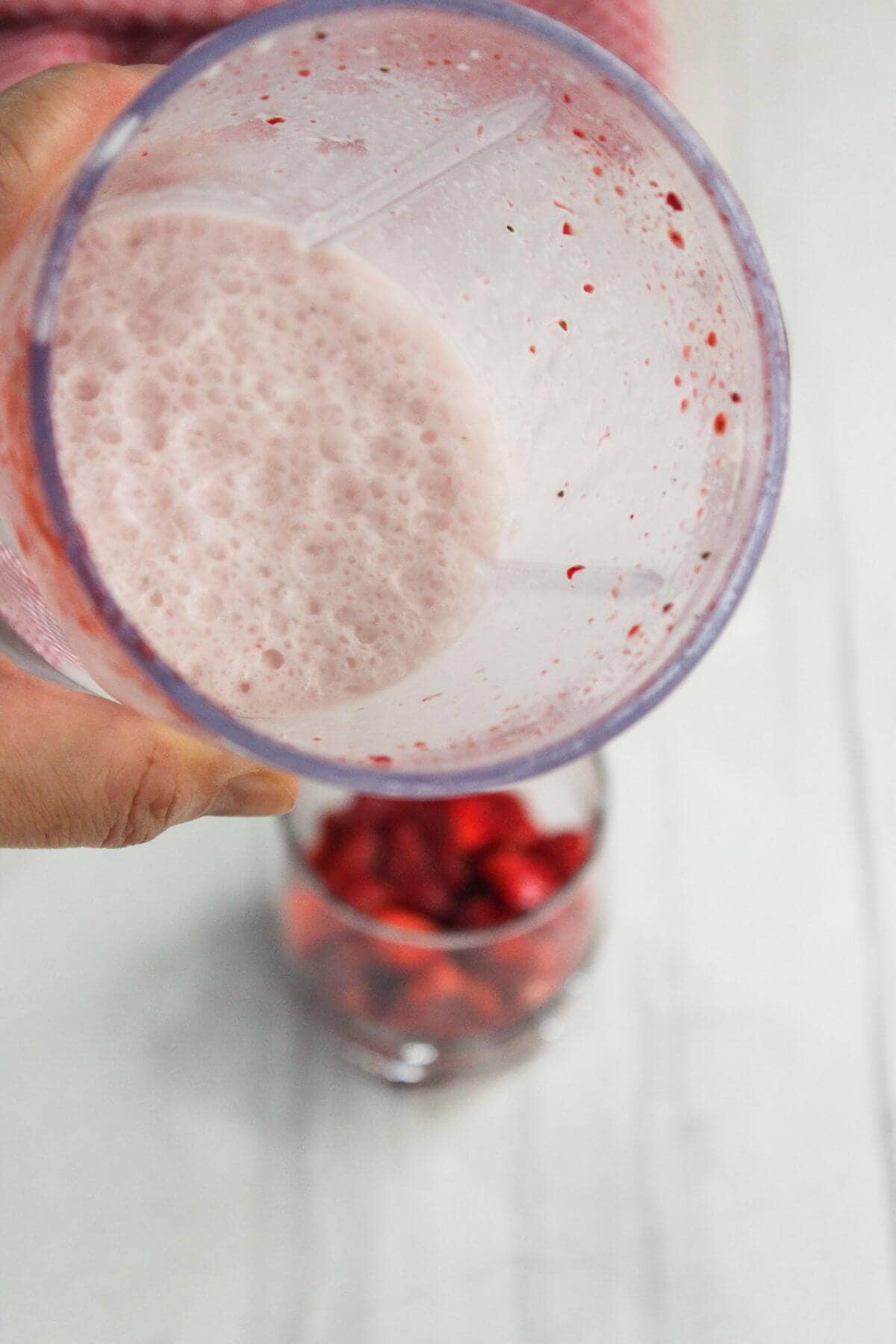 A hand holds a blender cup with pink frothy liquid above a glass filled with chopped strawberries on a white surface.