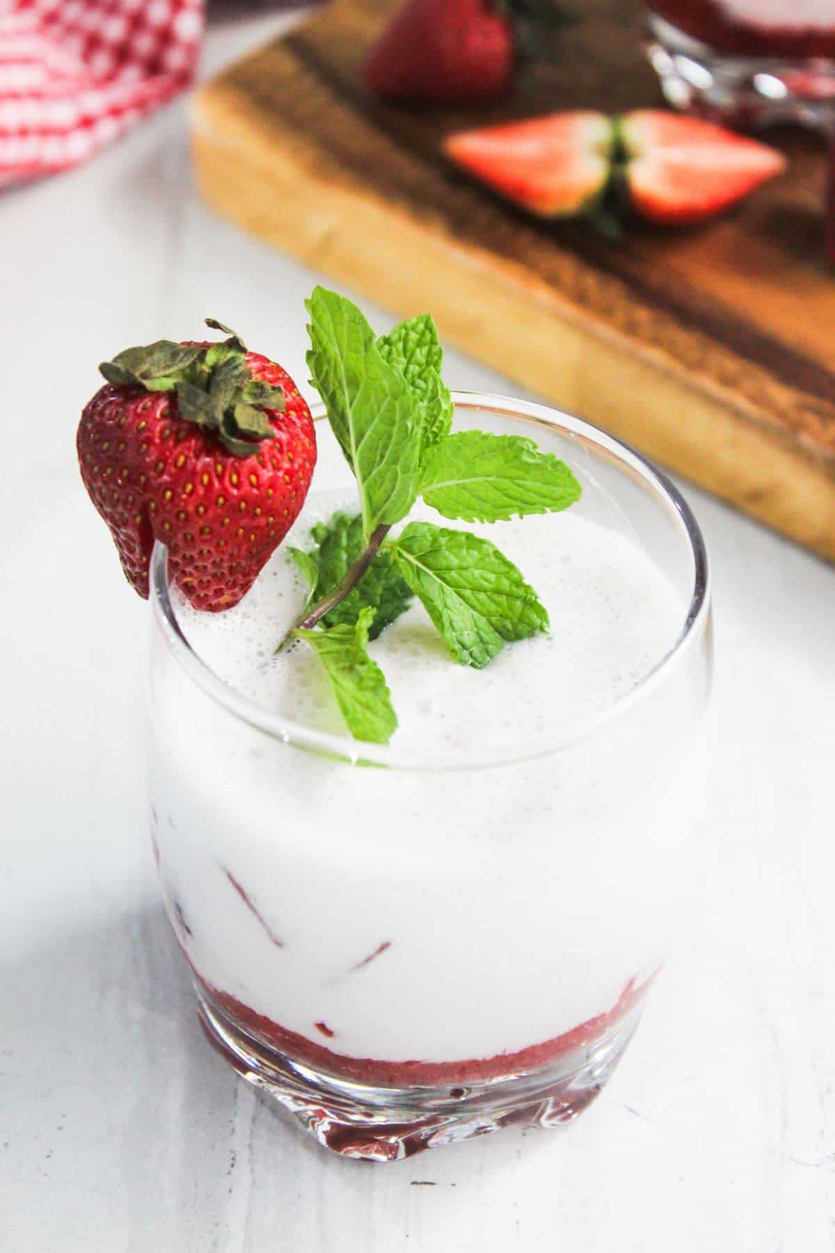 A glass of frothy white beverage with a layer of red puree at the bottom, garnished with a whole strawberry and fresh mint sprig. A wooden board with cut strawberries is in the background.