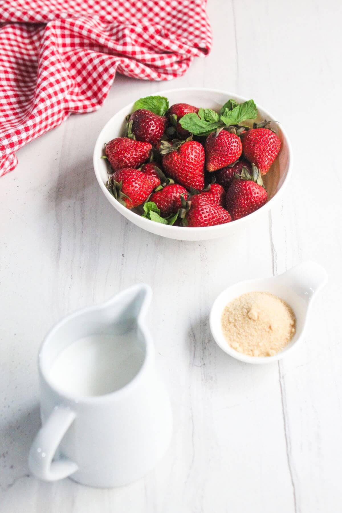 A bowl of fresh strawberries, a small white pitcher of milk, and a dish of granulated sugar on a white surface with a red checkered cloth.