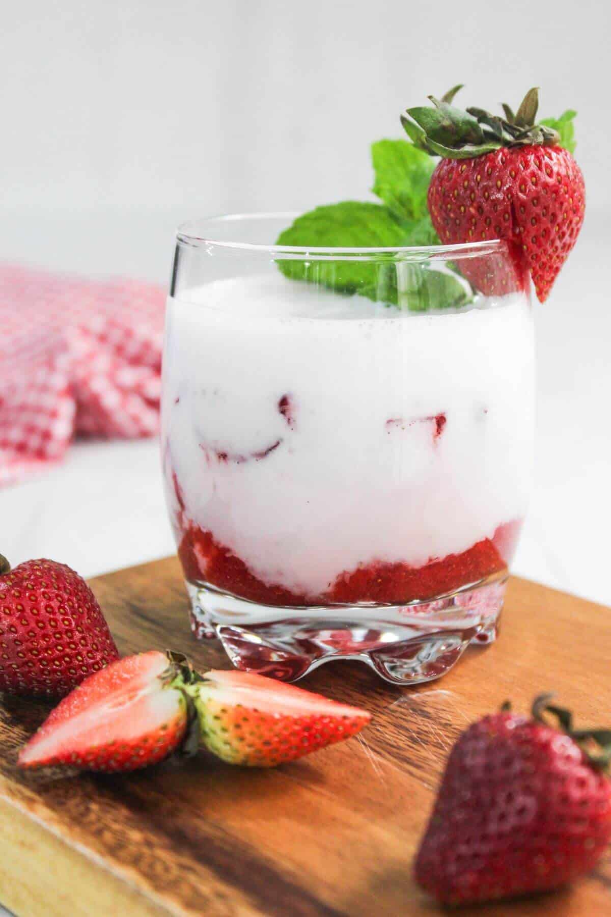 A glass of strawberry milk topped with a mint sprig and a whole strawberry, surrounded by fresh strawberries on a wooden board.