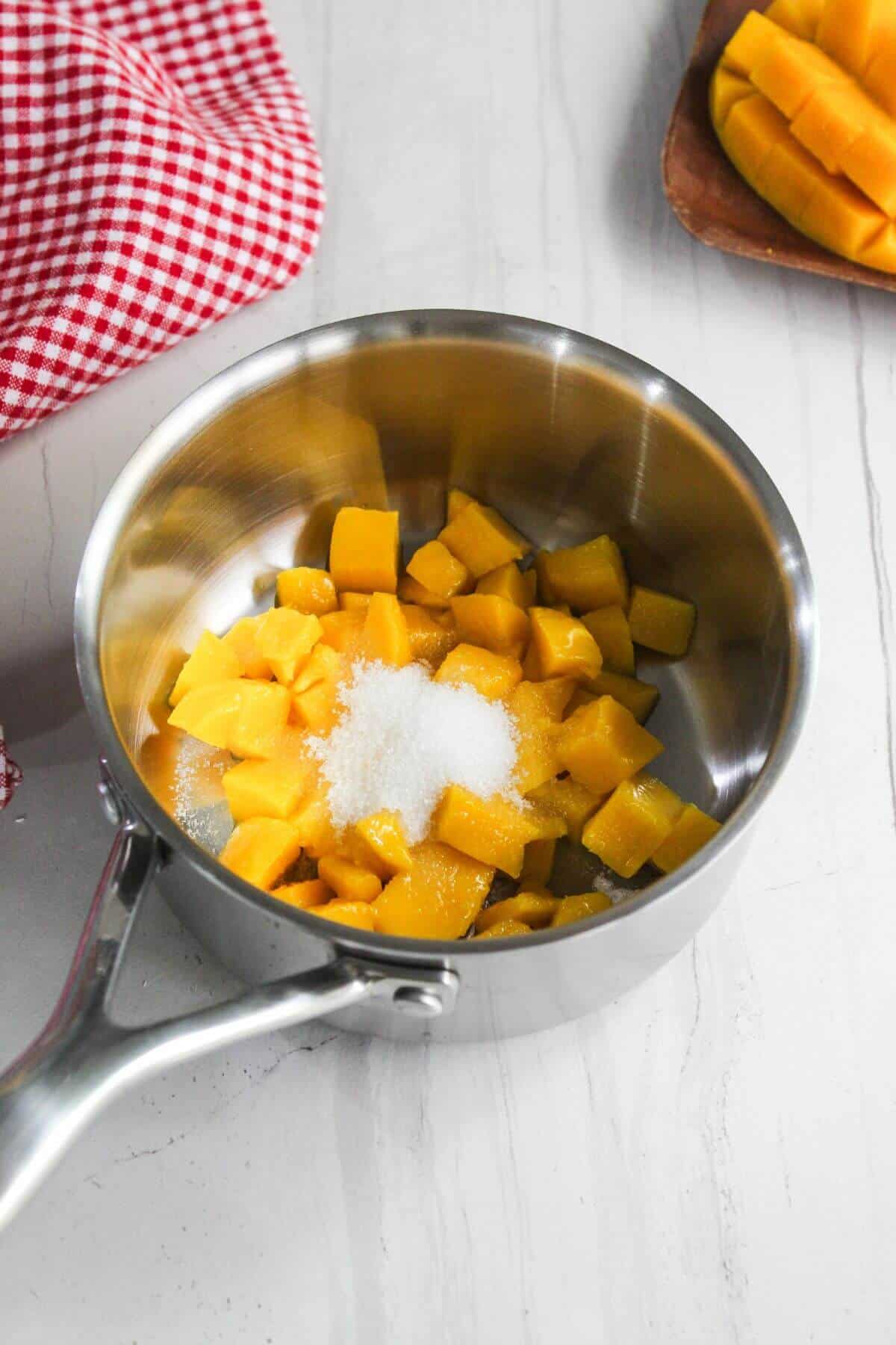 A stainless steel saucepan containing diced mango and a heap of granulated sugar sits on a white surface, with a red checkered cloth and a plate of more diced mango nearby.