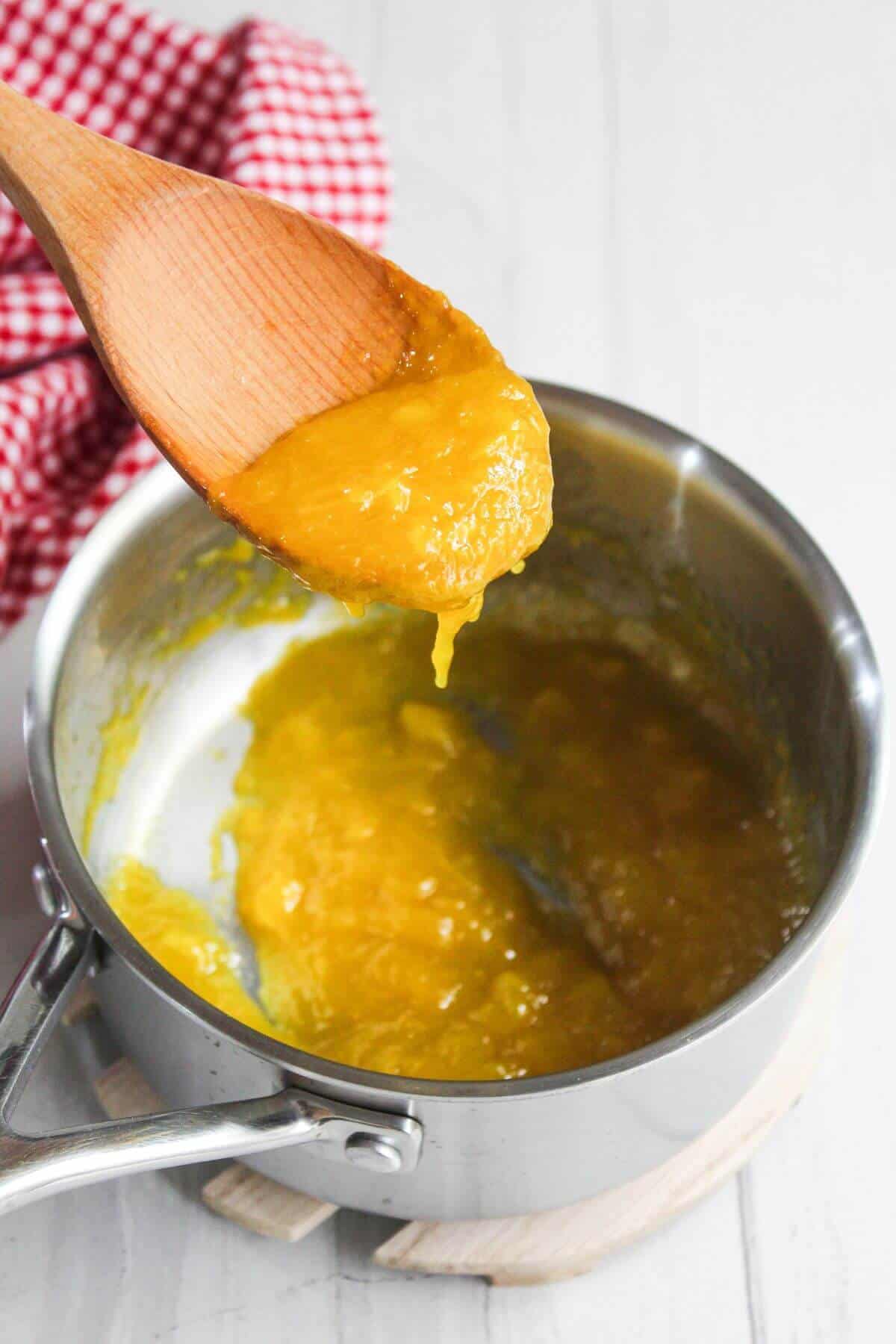 A wooden spoon holds a thick yellow mixture over a saucepan with more of the same mixture inside. A red and white checkered cloth is partially visible in the background.