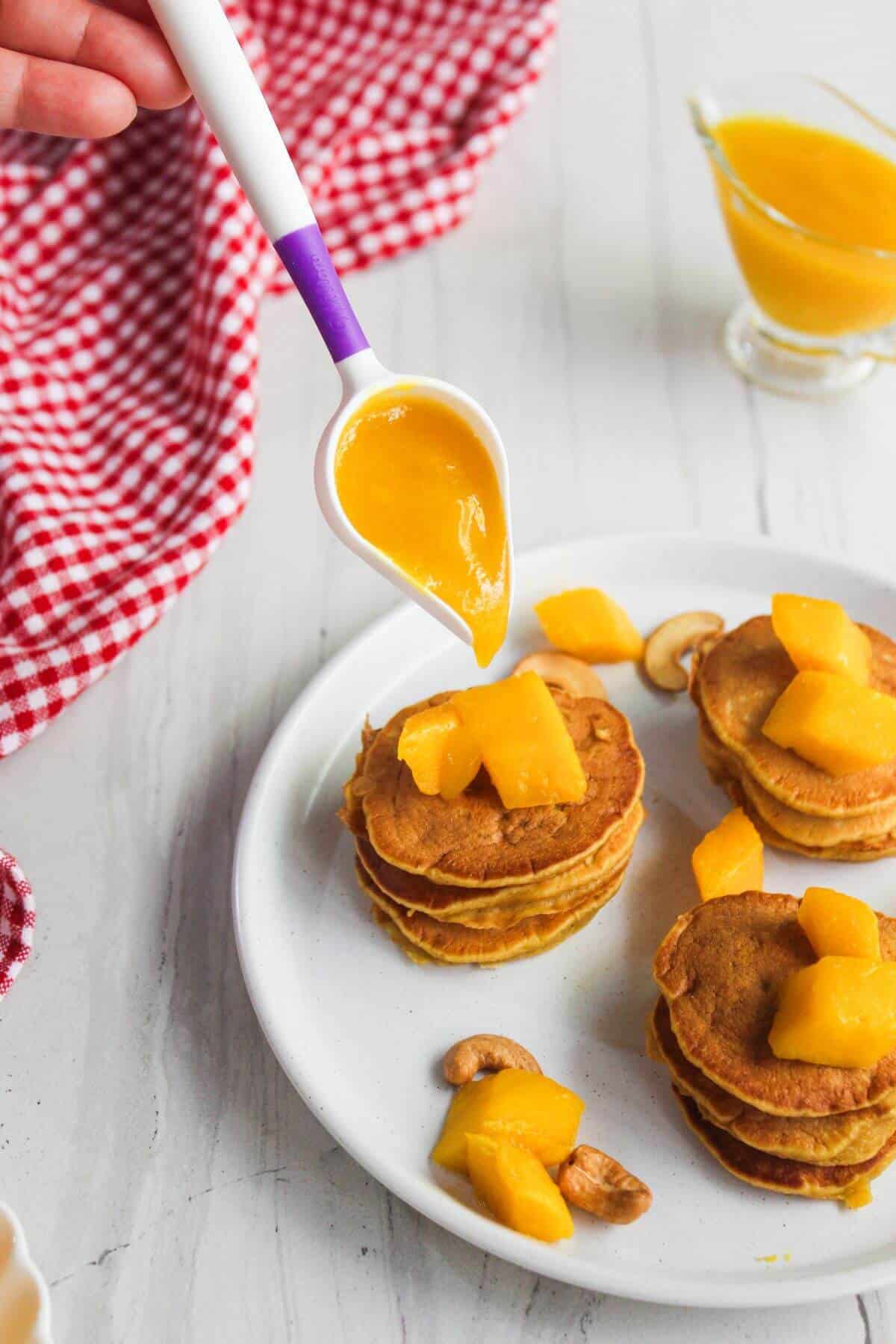 A hand pours mango sauce from a spoon onto a stack of pancakes topped with mango pieces and cashews, with a red checkered cloth nearby.