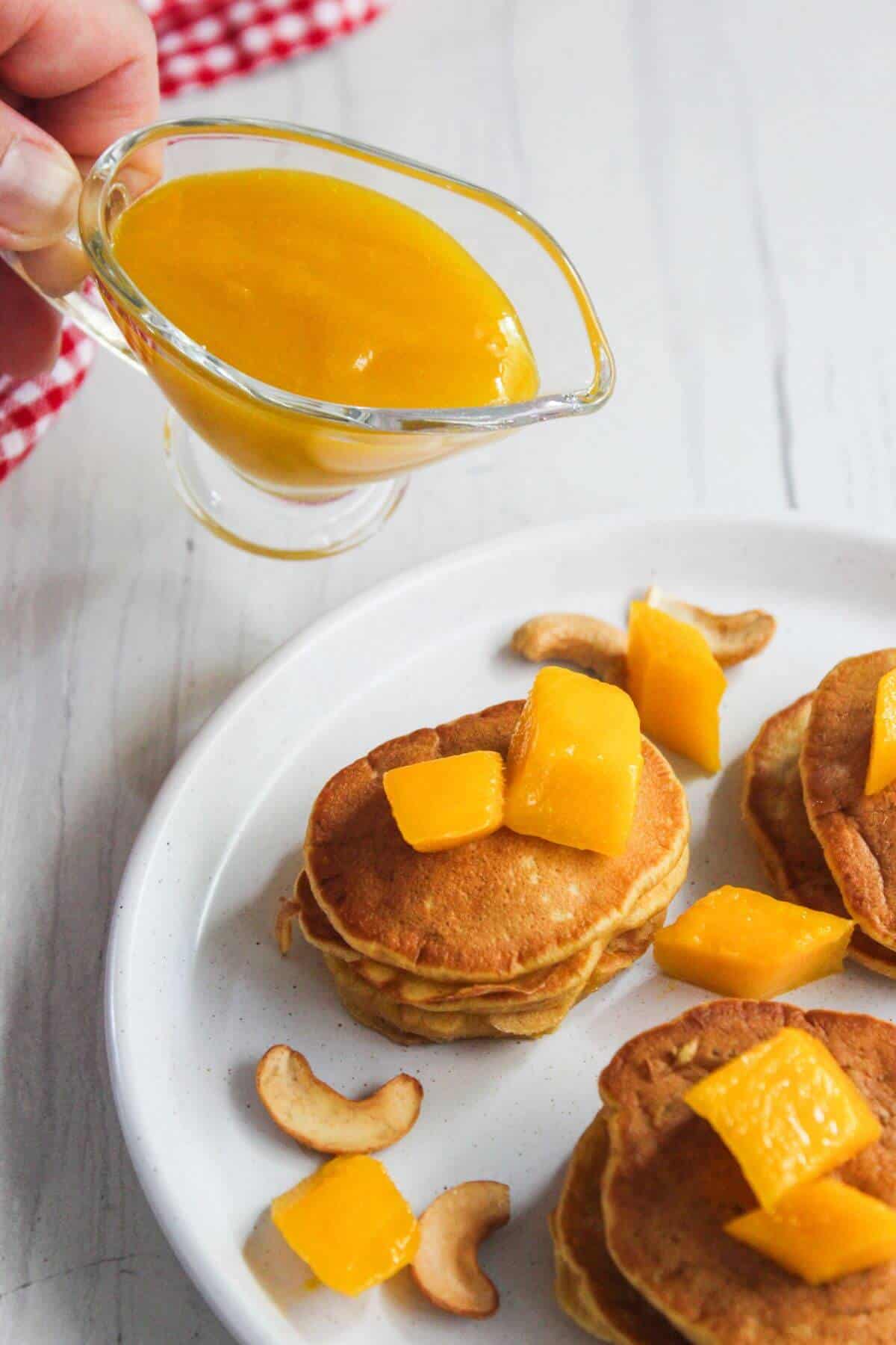 A hand pours mango sauce from a small glass pitcher onto a plate of mini pancakes topped with mango cubes and surrounded by cashew nuts.