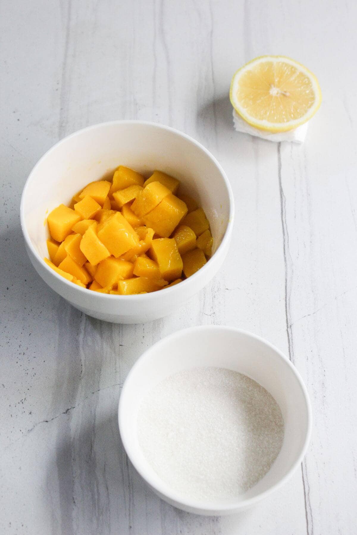 A bowl of diced mango, a bowl of granulated sugar, and half a lemon on a white marble surface.