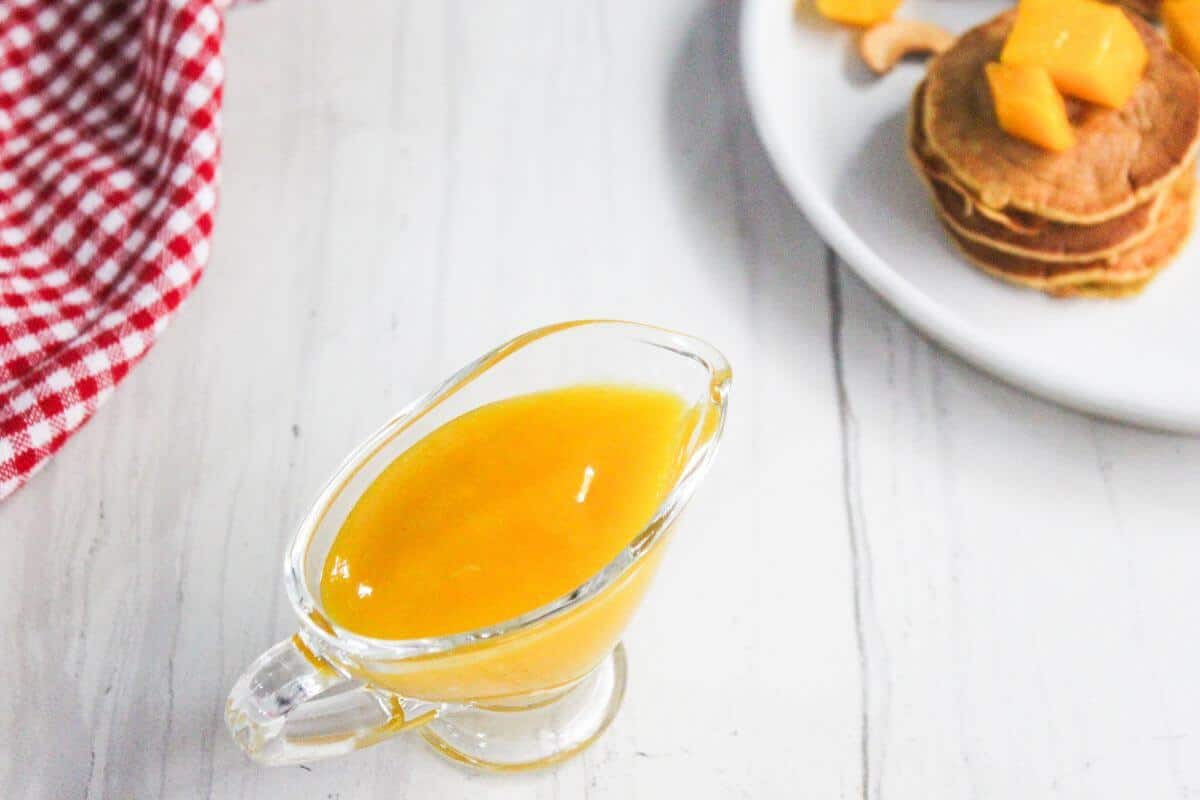 A glass gravy boat filled with mango sauce sits on a white surface next to a plate of pancakes topped with mango cubes and a red checkered cloth.