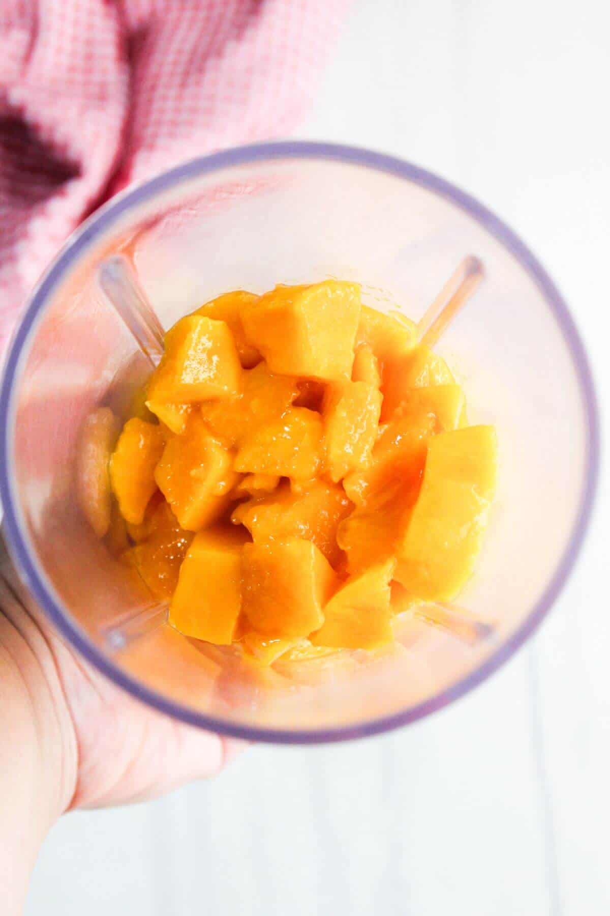 Chunks of mango inside a blender cup, ready to be blended, with a hand holding the cup and a red cloth visible in the background.