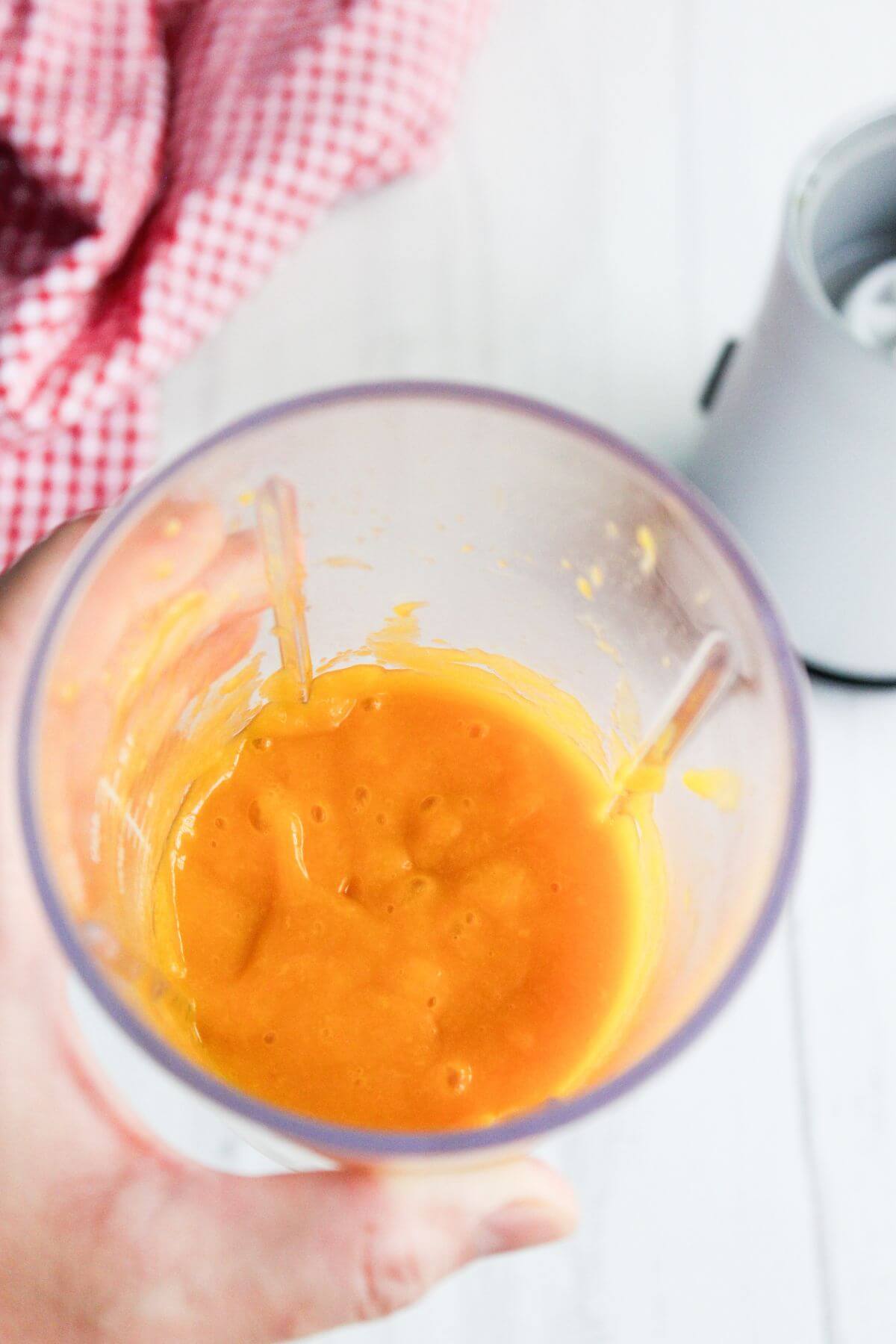 A hand holds a blender cup filled with thick orange puree, with a blender base and a red checkered cloth in the background.