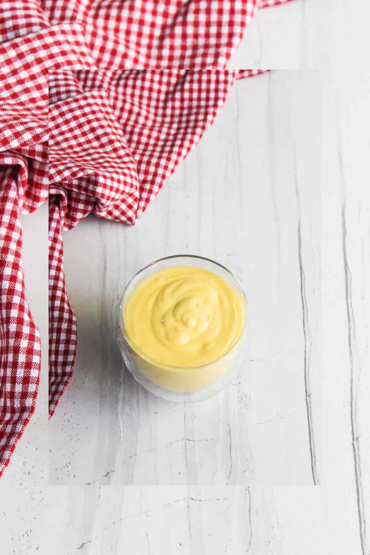 A small glass cup filled with yellow mayonnaise sits on a white marble surface next to a red and white checkered cloth.