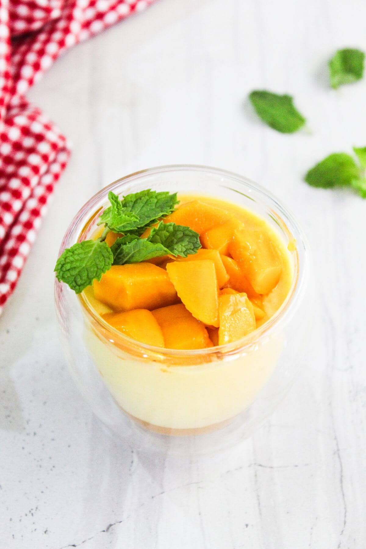 A clear glass cup filled with a creamy dessert topped with diced mango pieces and mint leaves sits on a white surface next to a red checkered cloth and mint sprigs.