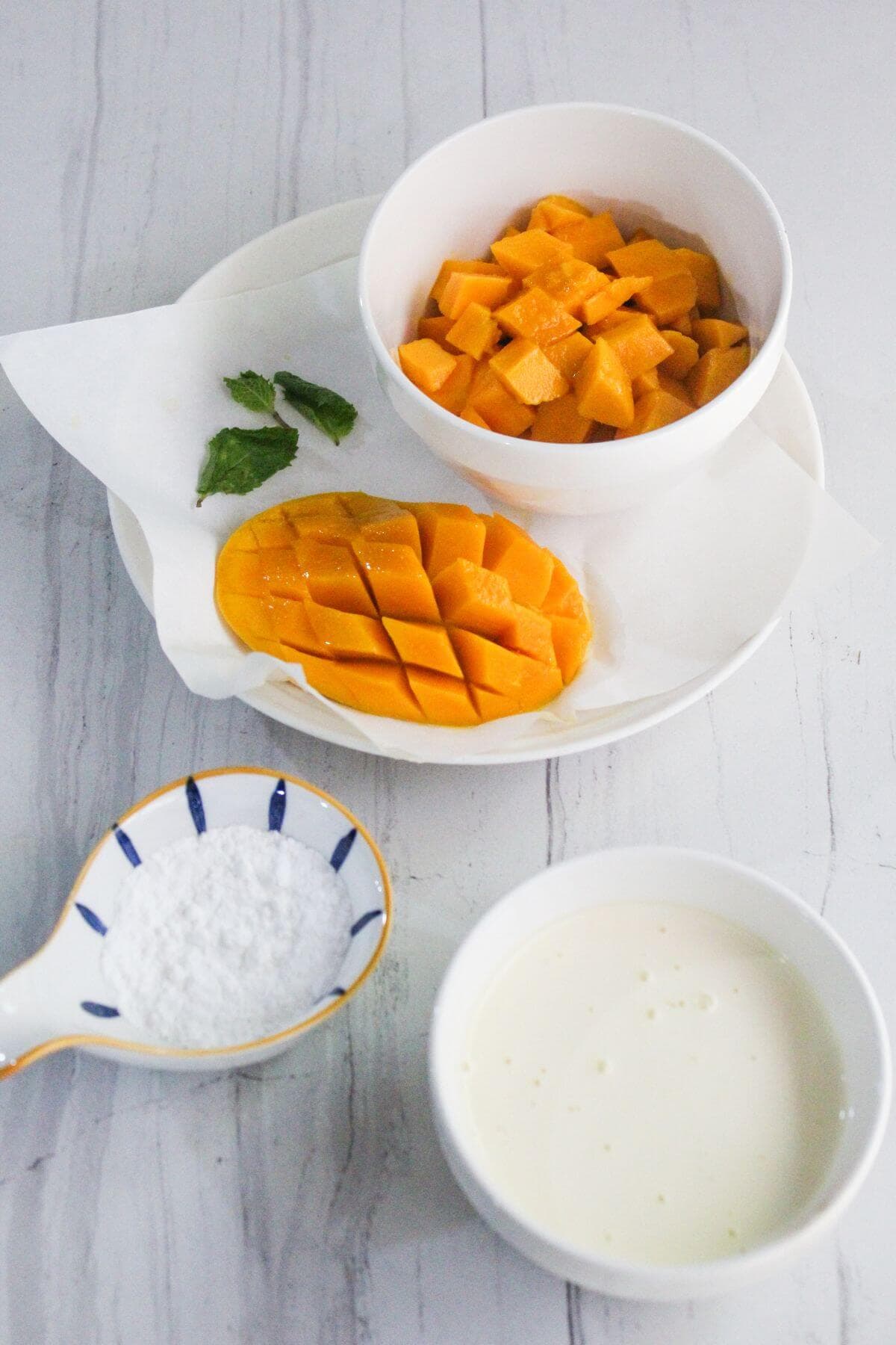 Bowls containing diced mango, sliced mango, powdered sugar, and cream are arranged on a white surface with a few mint leaves for garnish.
