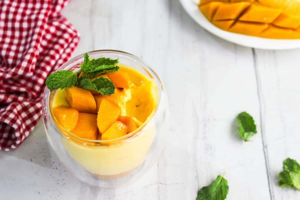 A glass cup of mango pudding topped with fresh mango cubes and mint leaves sits on a white table next to a red checkered cloth and a plate of sliced mango.