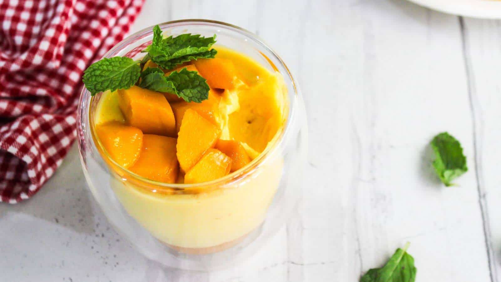 A glass cup of mango mousse topped with fresh mango cubes and mint leaves sits on a white table next to a red checkered cloth and a plate of sliced mango.