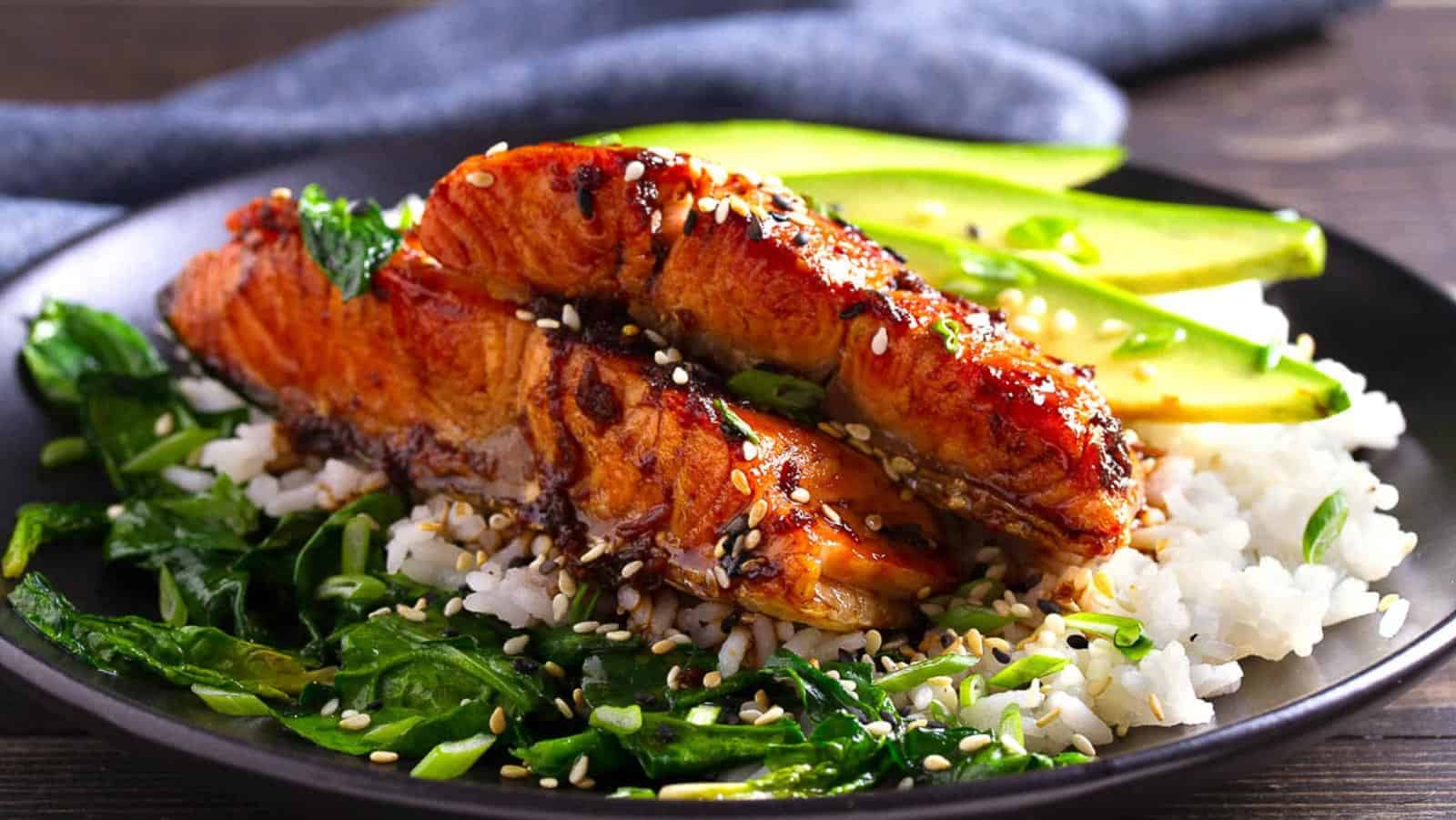 Grilled salmon fillet served on white rice with sautéed greens, avocado slices, and sprinkled sesame seeds on a black plate.
