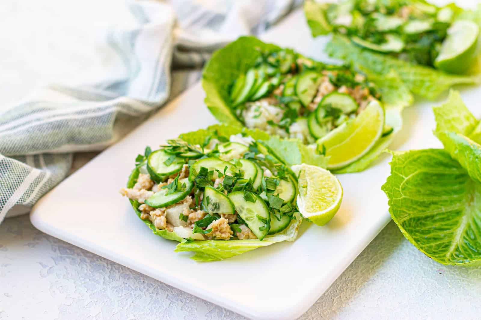Lettuce wraps filled with chopped chicken, sliced cucumber, herbs, and lime wedges on a white rectangular plate.