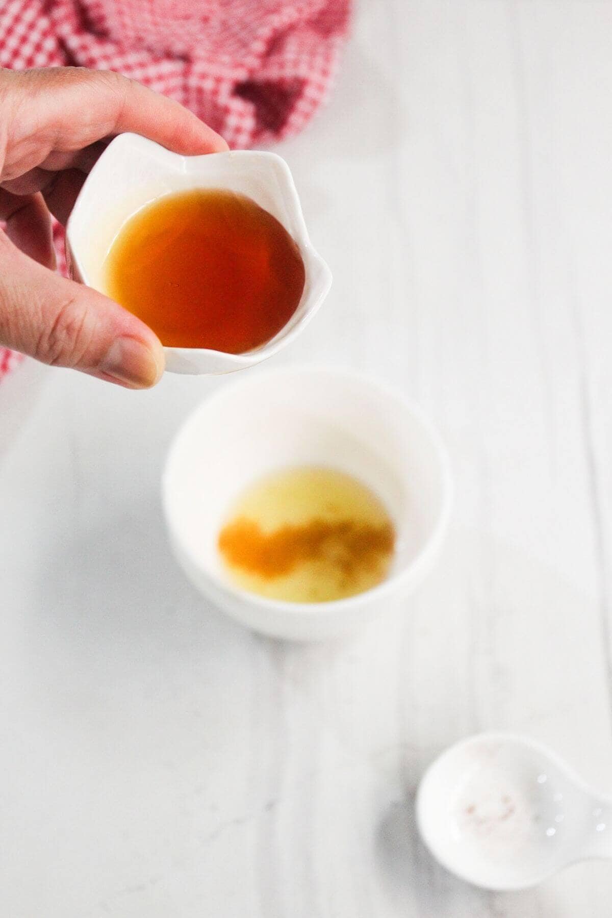 A hand holds a small white bowl with amber liquid above a larger white bowl containing a mixture, on a white surface with a red cloth in the background.