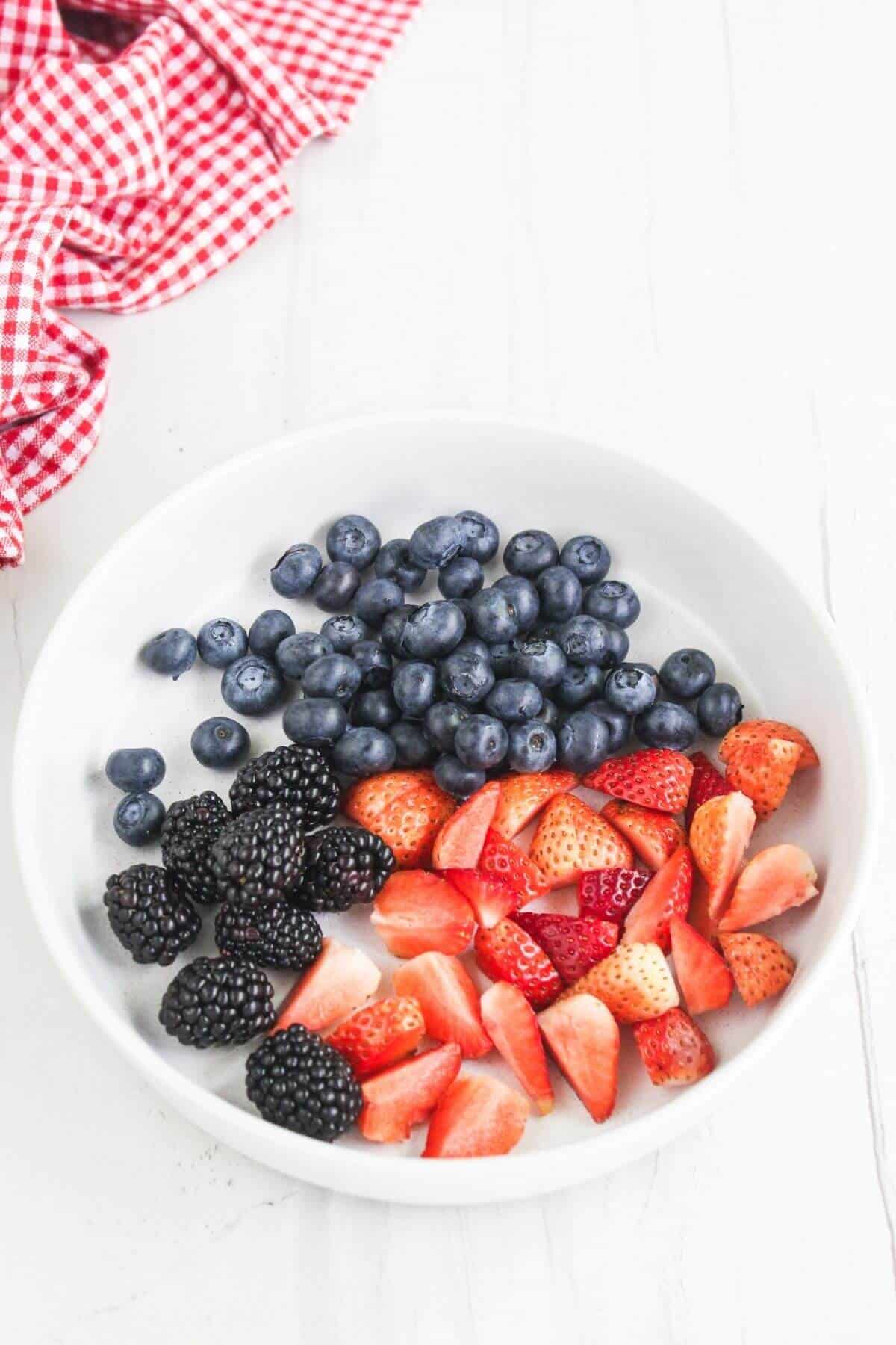 A white bowl containing fresh blueberries, strawberries, and blackberries, placed on a white surface with a red checkered cloth nearby.