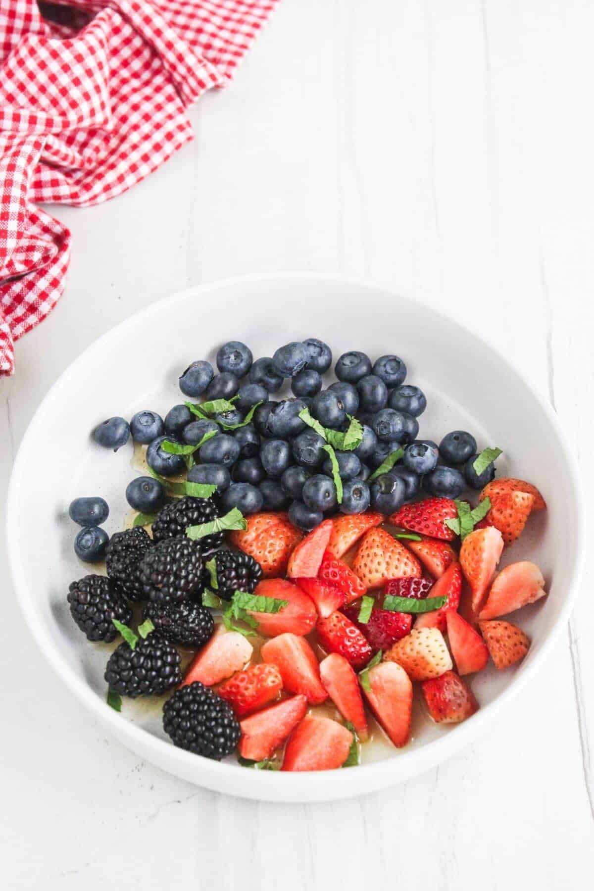 A white bowl filled with blueberries, sliced strawberries, and blackberries, garnished with chopped mint, sits on a white surface next to a red and white checkered cloth.