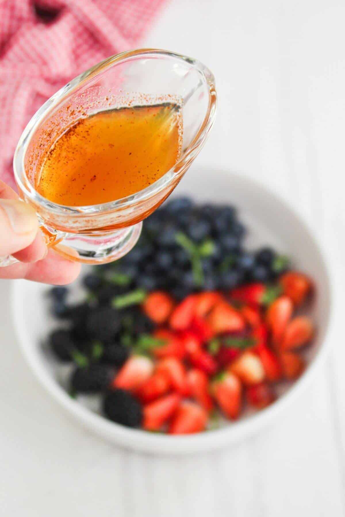 A hand holds a small glass pitcher of honey vinaigrette above a bowl filled with blueberries, strawberries, and blackberries.