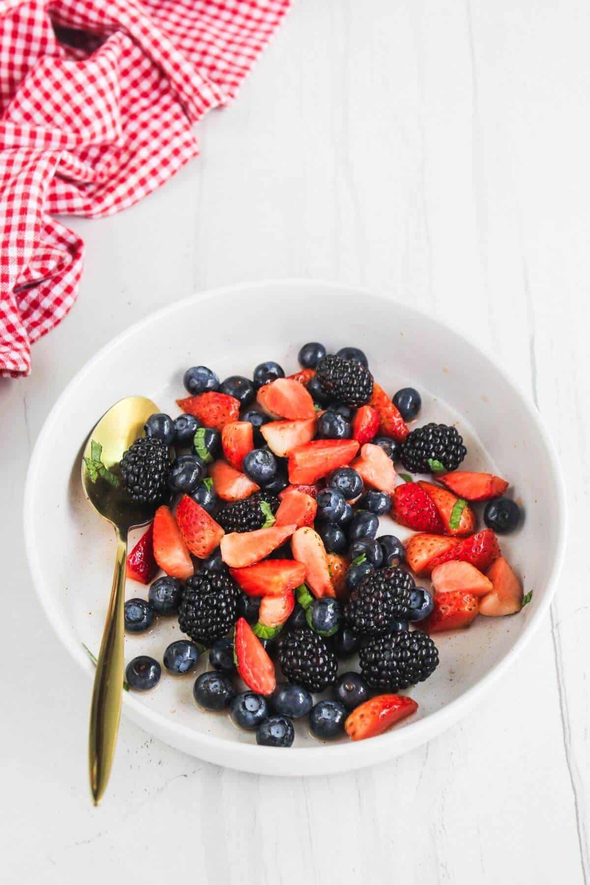 A white bowl filled with sliced strawberries, blackberries, blueberries, and fresh mint sits on a white surface with a gold spoon and a red checkered cloth nearby.
