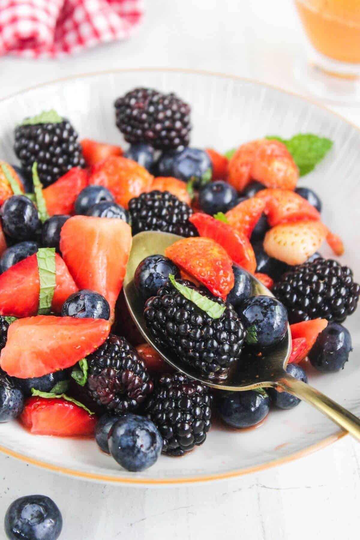 A bowl of mixed fresh berries, including strawberries, blackberries, and blueberries, garnished with mint leaves. A spoon rests inside the bowl.