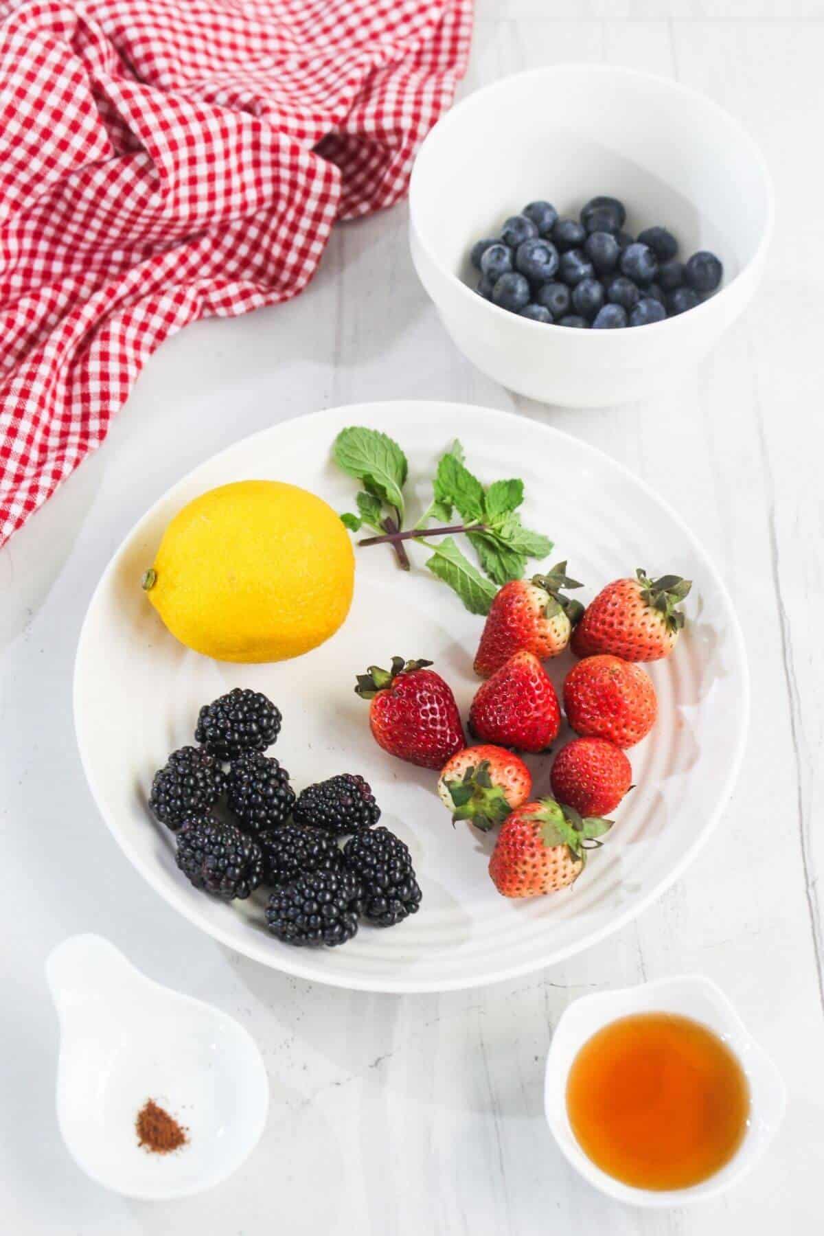 A white plate with strawberries, blackberries, a lemon, and mint leaves. Nearby are bowls of blueberries, ground spice, and a small bowl of liquid, on a white surface with a red checkered cloth.