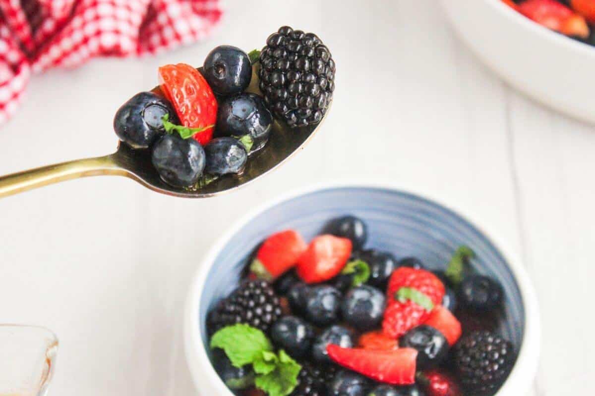A spoonful of mixed berries, including blackberries, blueberries, and sliced strawberries, held above a bowl of similar fruit salad on a white surface.