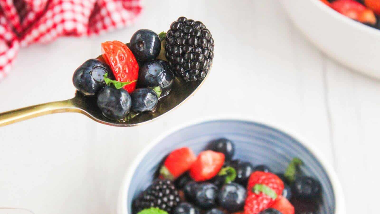 A spoon holding blueberries, a sliced strawberry, and a blackberry above a bowl of mixed berries on a white surface.