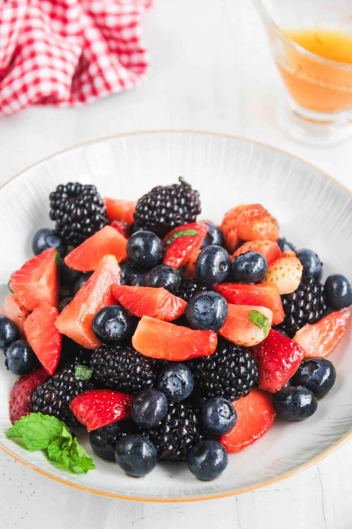 A bowl filled with fresh strawberries, blackberries, and blueberries, garnished with mint, sits on a white surface next to a glass cup of liquid and a red checkered cloth.
