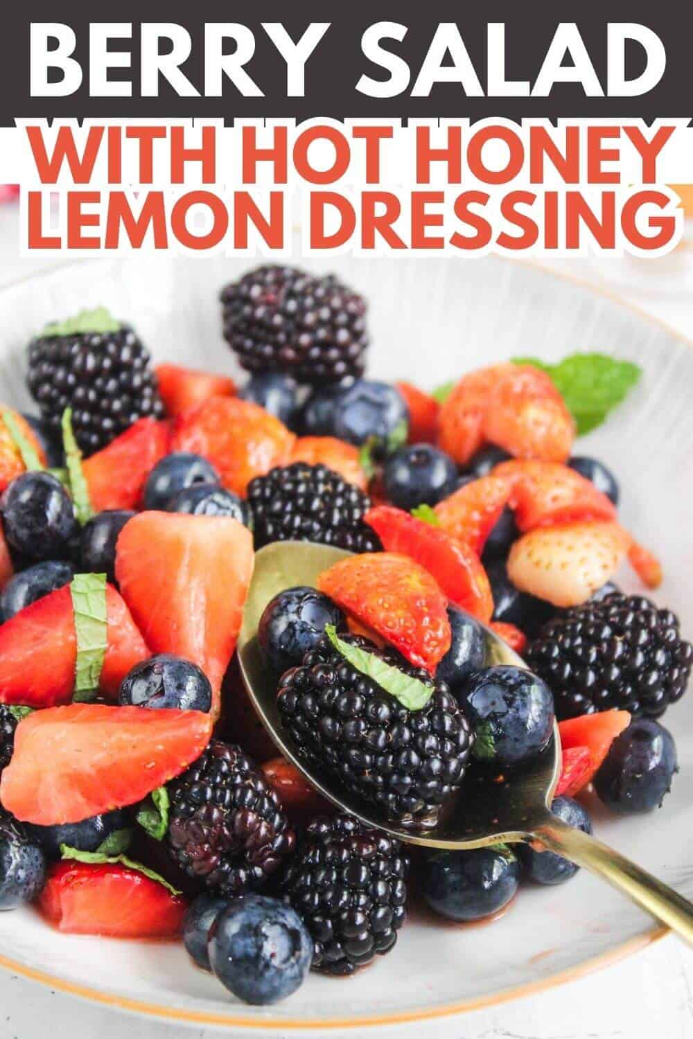A bowl of mixed berries including strawberries, blackberries, and blueberries with a spoon, garnished with mint leaves. Text above reads "Berry Salad with Hot Honey Lemon Dressing.