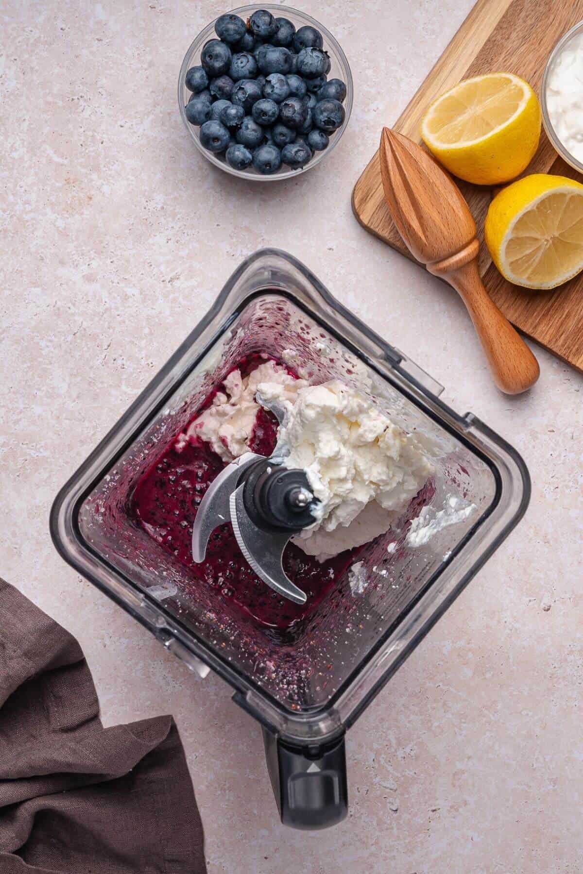 Overhead view of a blender with blueberries and cream cheese inside, next to fresh blueberries in a bowl, halved lemons, and a wooden citrus juicer on a cutting board.