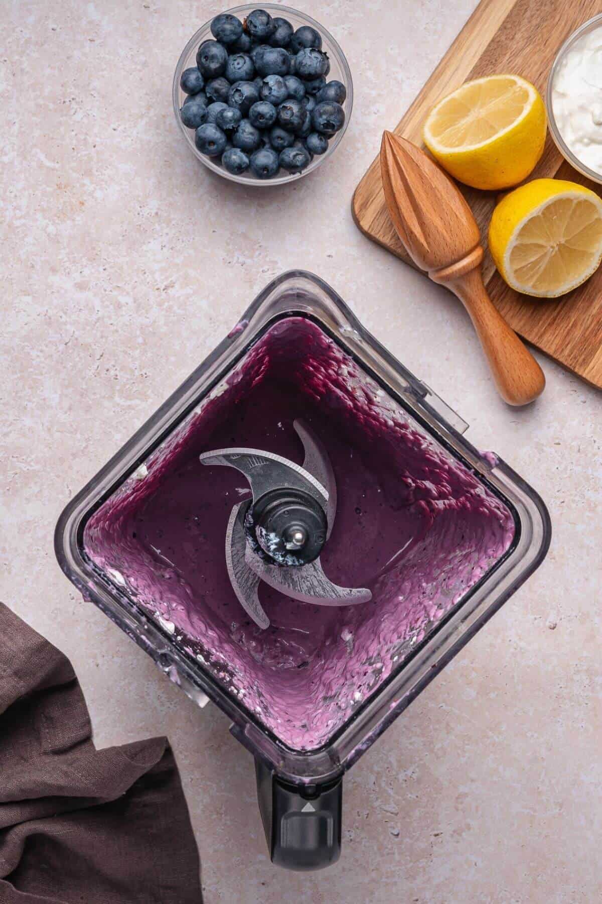 Top-down view of a blender with blueberry smoothie remnants, next to a bowl of blueberries, halved lemons, a wooden citrus juicer, and a small bowl of yogurt on a countertop.