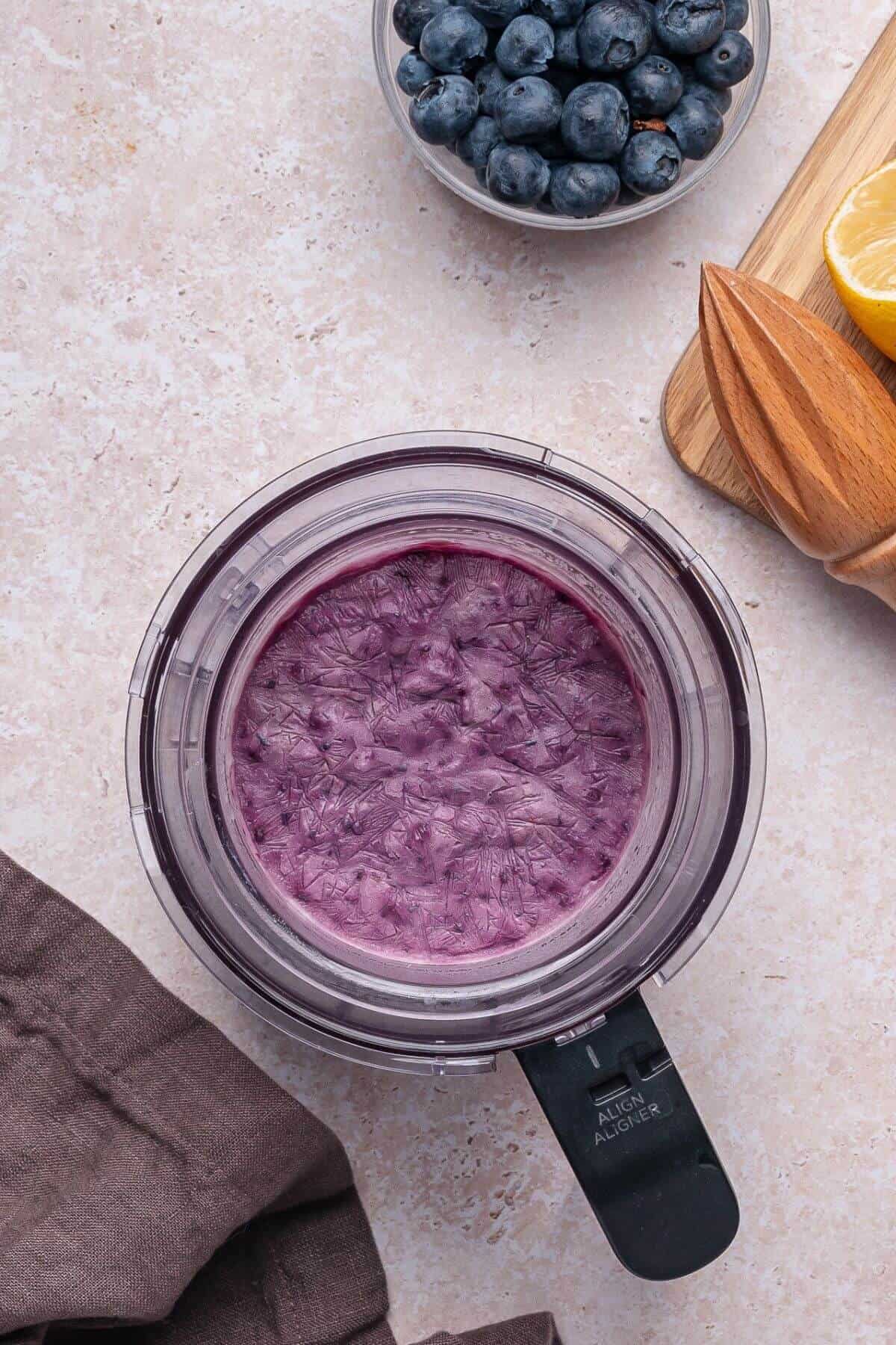 A blender containing a purple smoothie mixture sits on a countertop next to fresh blueberries, a lemon wedge, and a wooden citrus reamer.