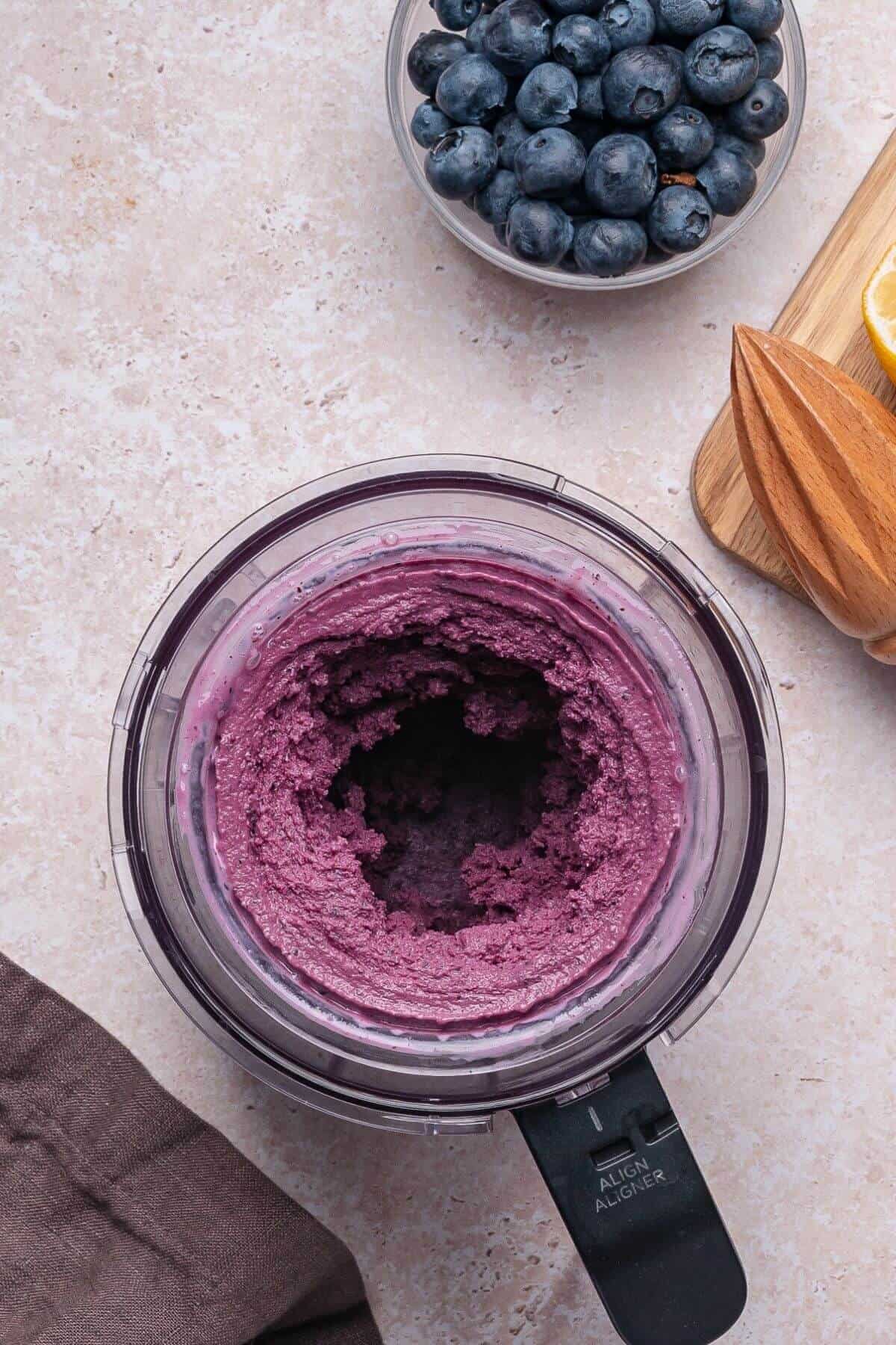 A food processor containing a purple blueberry mixture sits on a countertop near a bowl of blueberries, a wooden citrus juicer, and a lemon wedge.