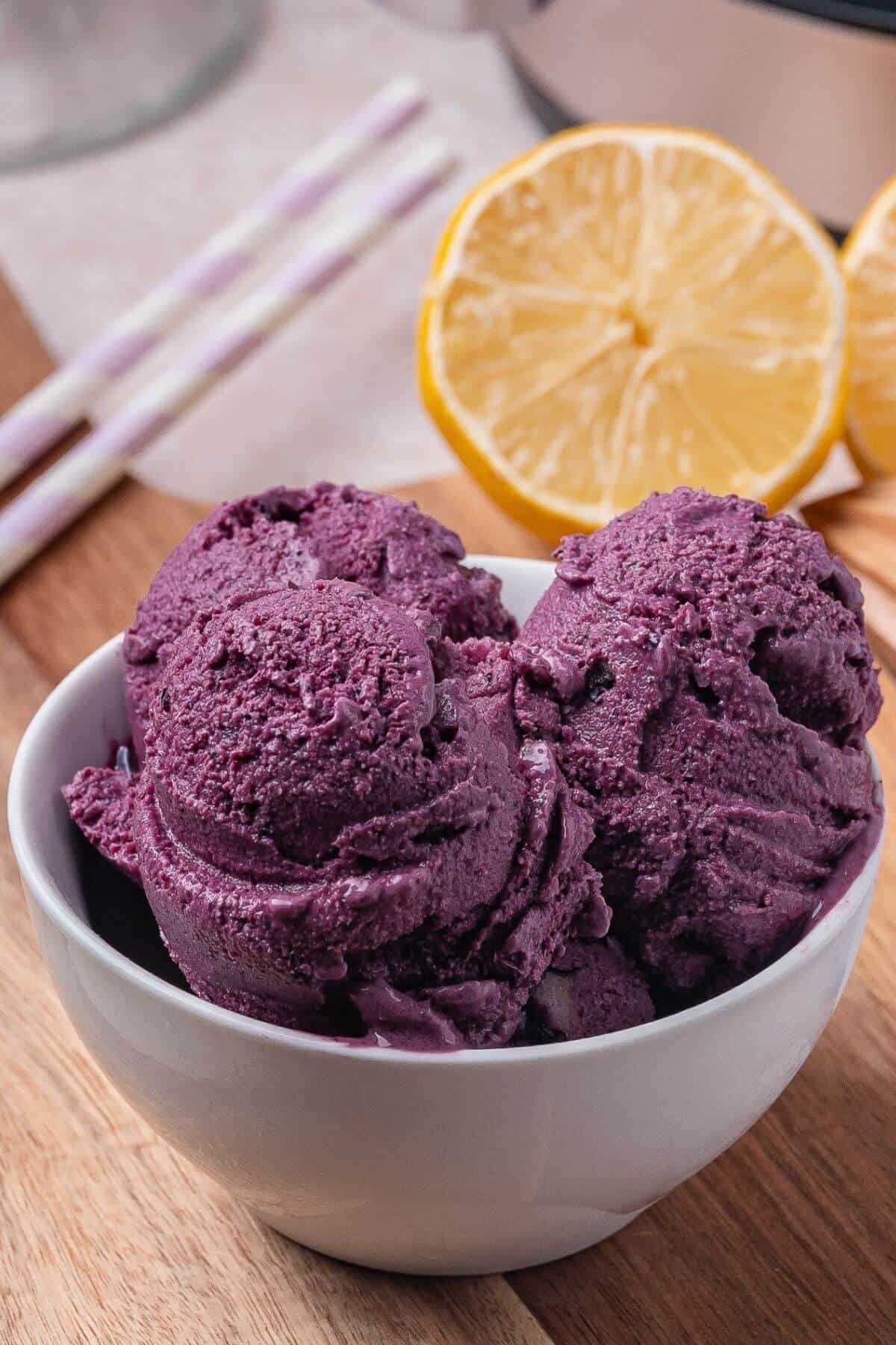 A white bowl filled with three scoops of dark purple ice cream sits on a wooden surface, with a halved lemon and striped straws in the background.