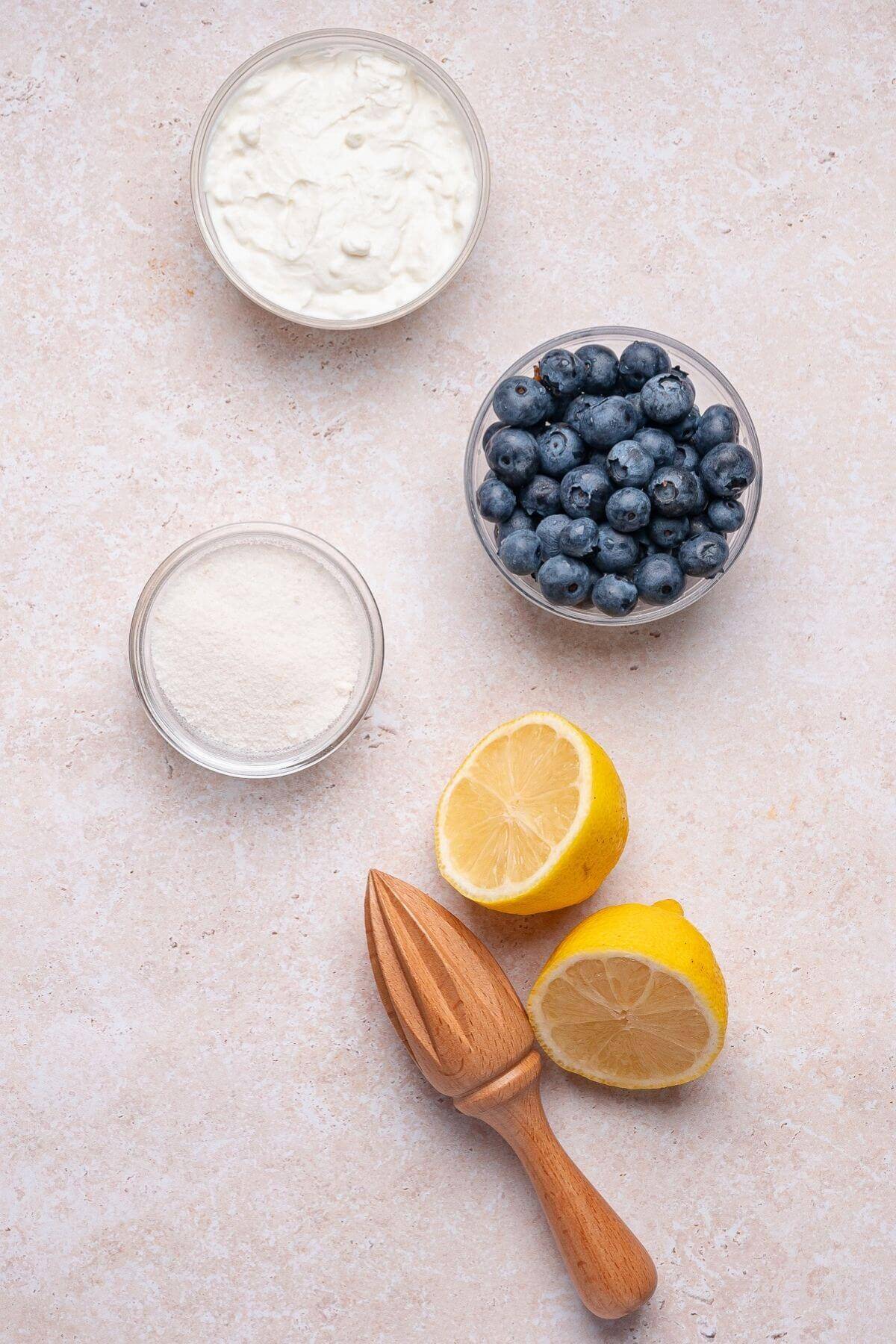 Three bowls containing yogurt, blueberries, and powder, plus two lemon halves and a wooden citrus reamer, arranged on a light countertop.