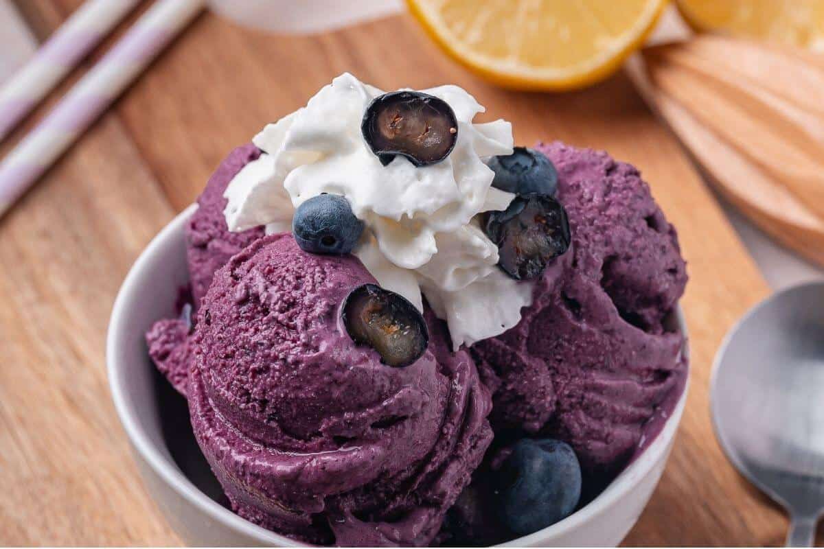 A bowl of purple blueberry ice cream topped with whipped cream and fresh blueberries, with lemon slices in the background.