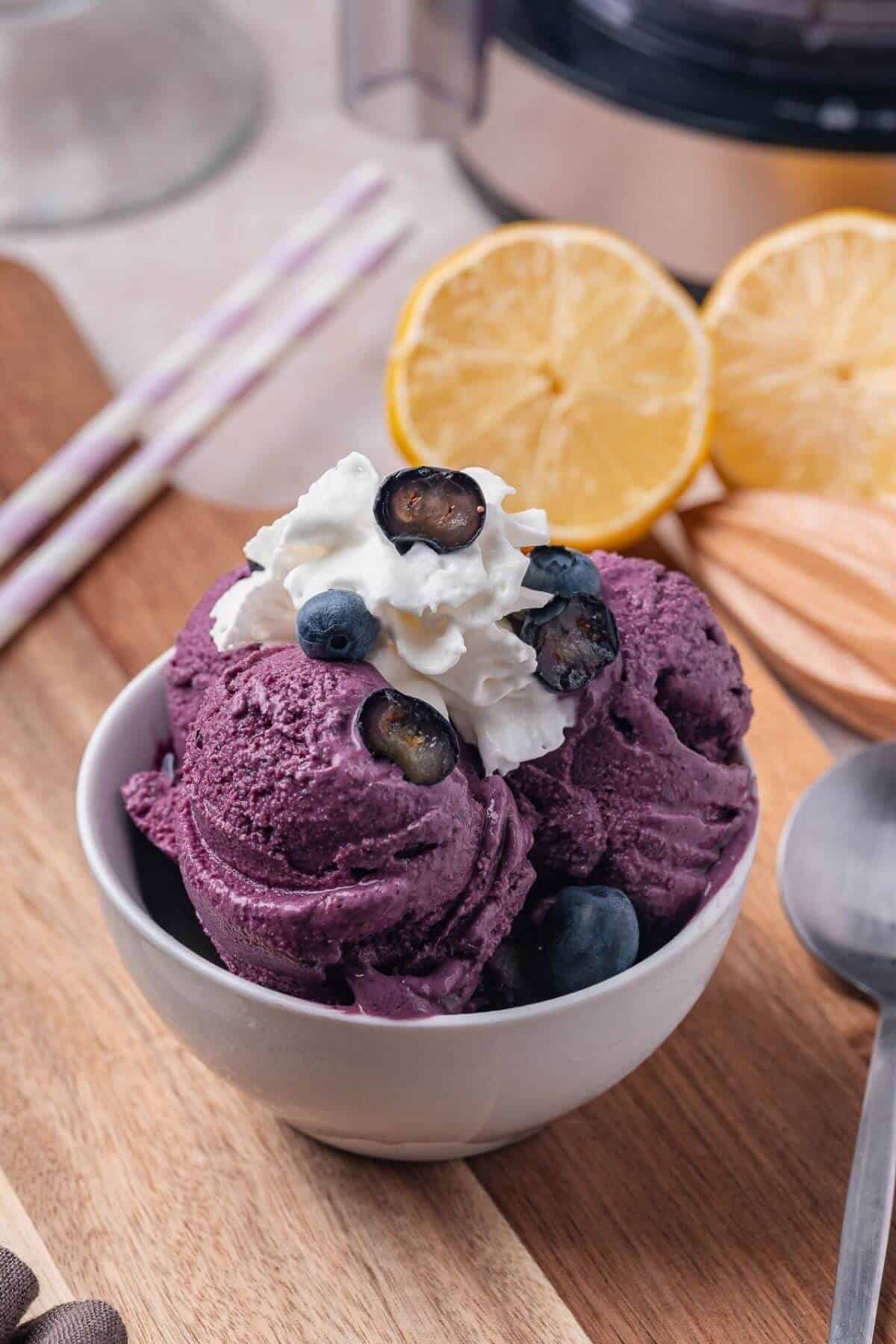 A bowl of purple blueberry ice cream topped with whipped cream and fresh blueberries, with sliced lemons and striped straws in the background.